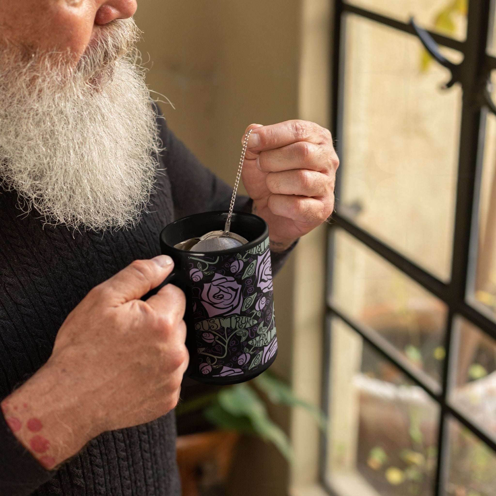 Older man with grey beard steeping tea in anti-Trump hidden message mug by a window