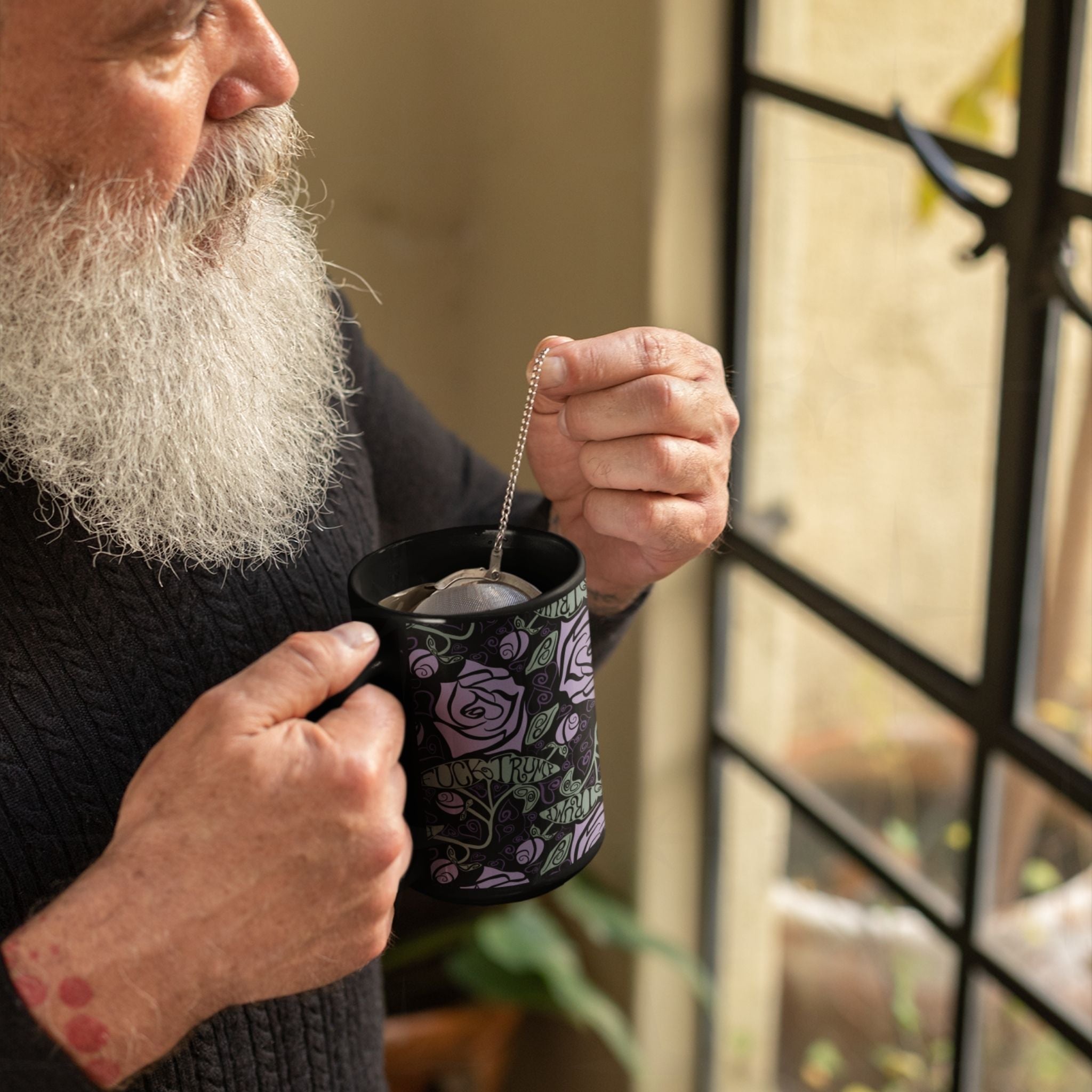 Older man with grey beard steeping tea in fuck Trump hidden message mug by a window