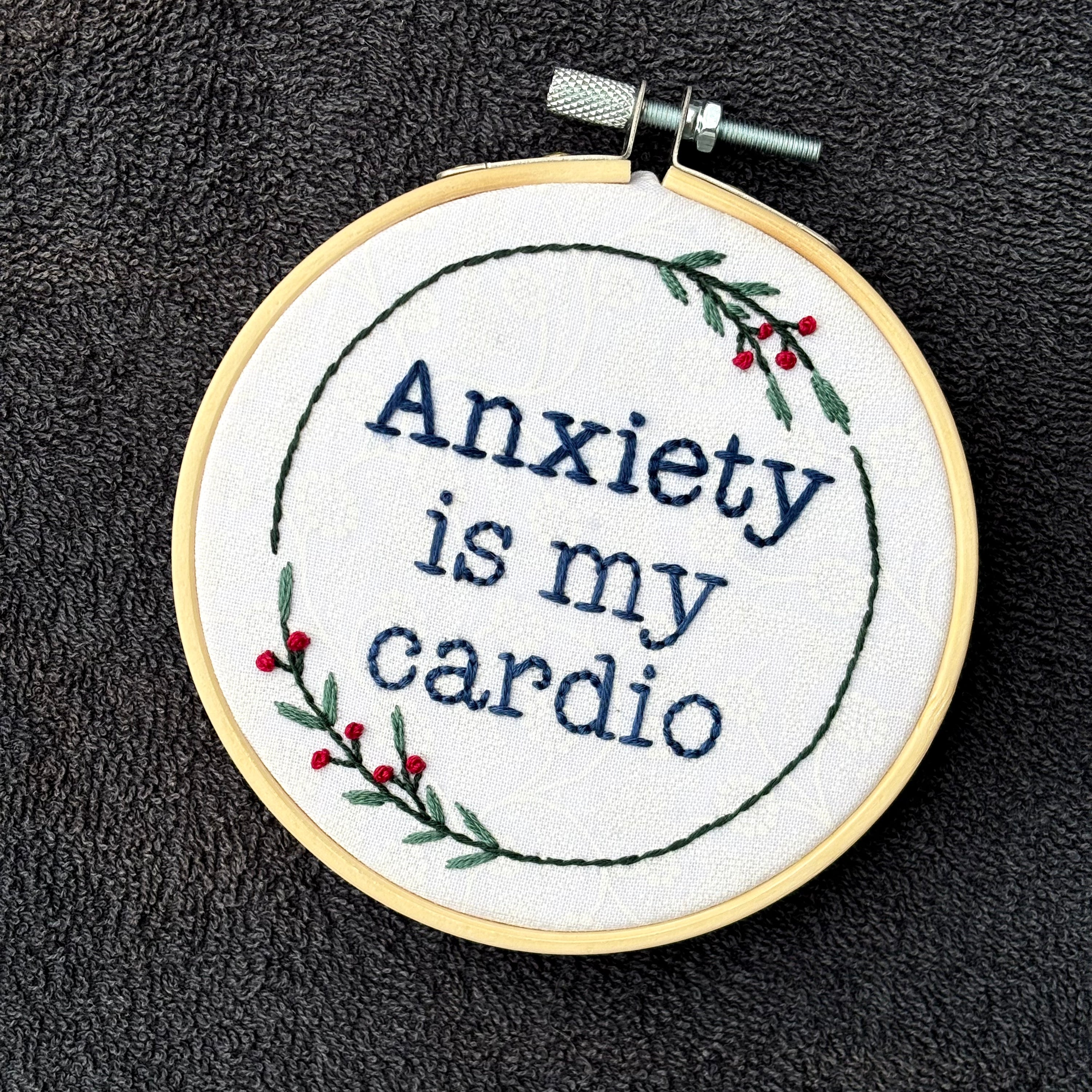 "Anxiety is my cardio." Quote hand embroidered in dark blue in typewriter font. Text is surrounded by minimalist floral wreath of dark green stems, green leaves, and red buds.. Fabric is white-on-white cotton. Displayed as wall art in a light wooden embroidery hoop.