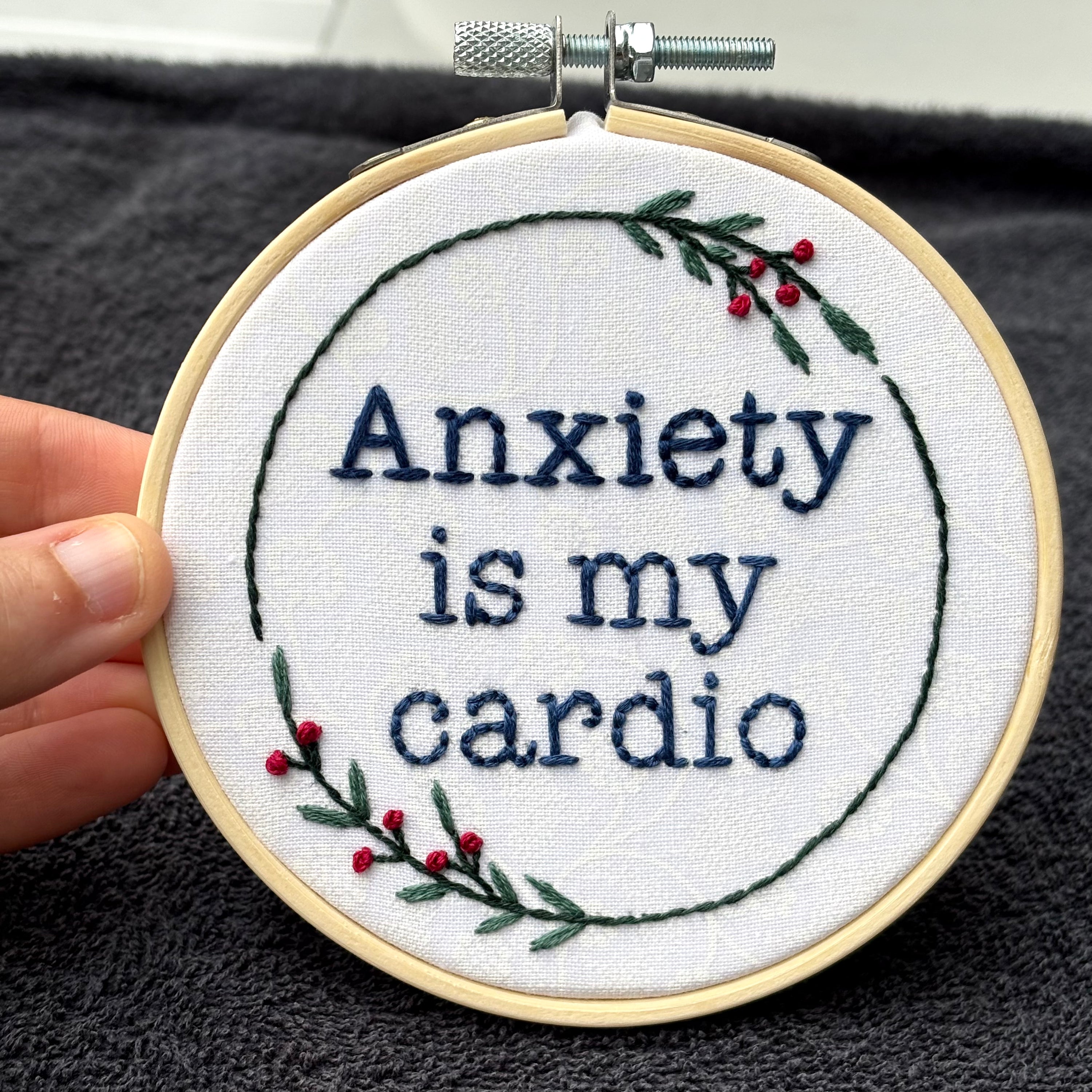 "Anxiety is my cardio." Quote hand embroidered in dark blue in typewriter font. Text is surrounded by minimalist floral wreath of dark green stems, green leaves, and red buds.. Fabric is white-on-white cotton. Displayed as wall art in a light wooden embroidery hoop.
