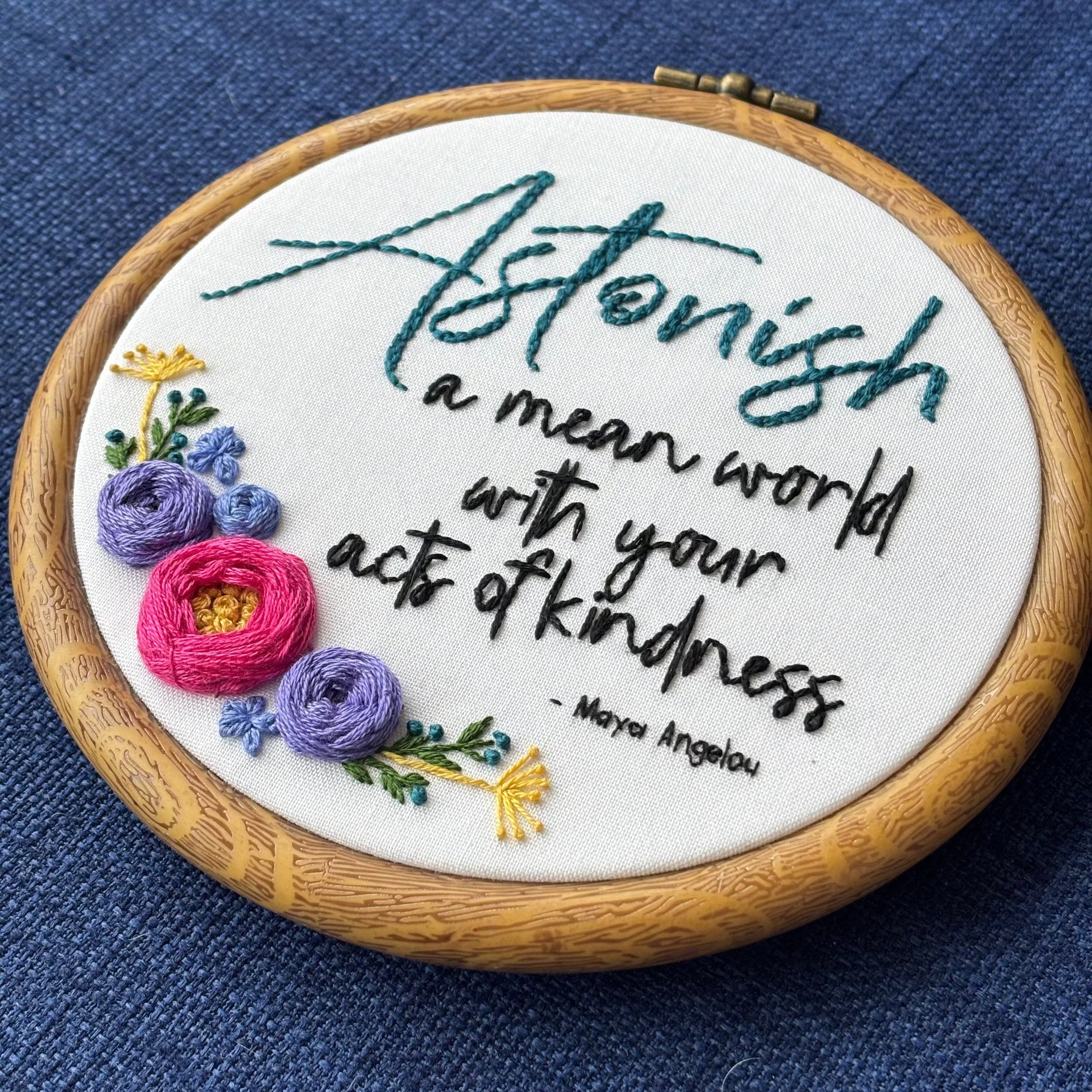 "Astonish a mean world with your acts of kindness. Maya Angelou." Astonish hand embroidered in dark teal. Remaining quote and attribution hand embroidered in black. In the lower left side, a cluster of flowers with bright pink and purple roses, golden pointy flowers, and green stems with leaves and dark teal flower buds. Art is displayed in a medium dark grained wood-look embroidery hoop.