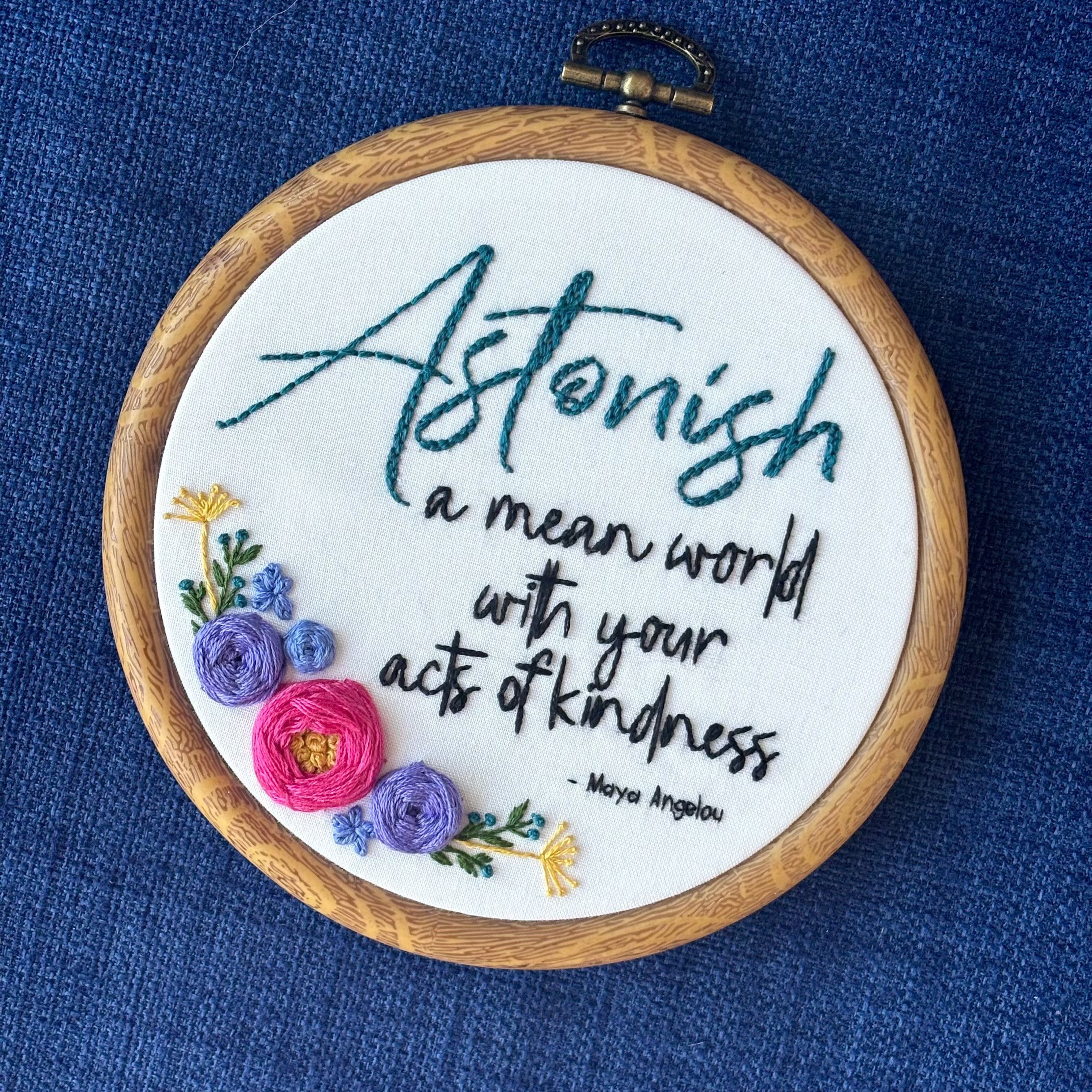 "Astonish a mean world with your acts of kindness. Maya Angelou." Astonish hand embroidered in dark teal. Remaining quote and attribution hand embroidered in black. In the lower left side, a cluster of flowers with bright pink and purple roses, golden pointy flowers, and green stems with leaves and dark teal flower buds. Art is displayed in a medium dark grained wood-look embroidery hoop.