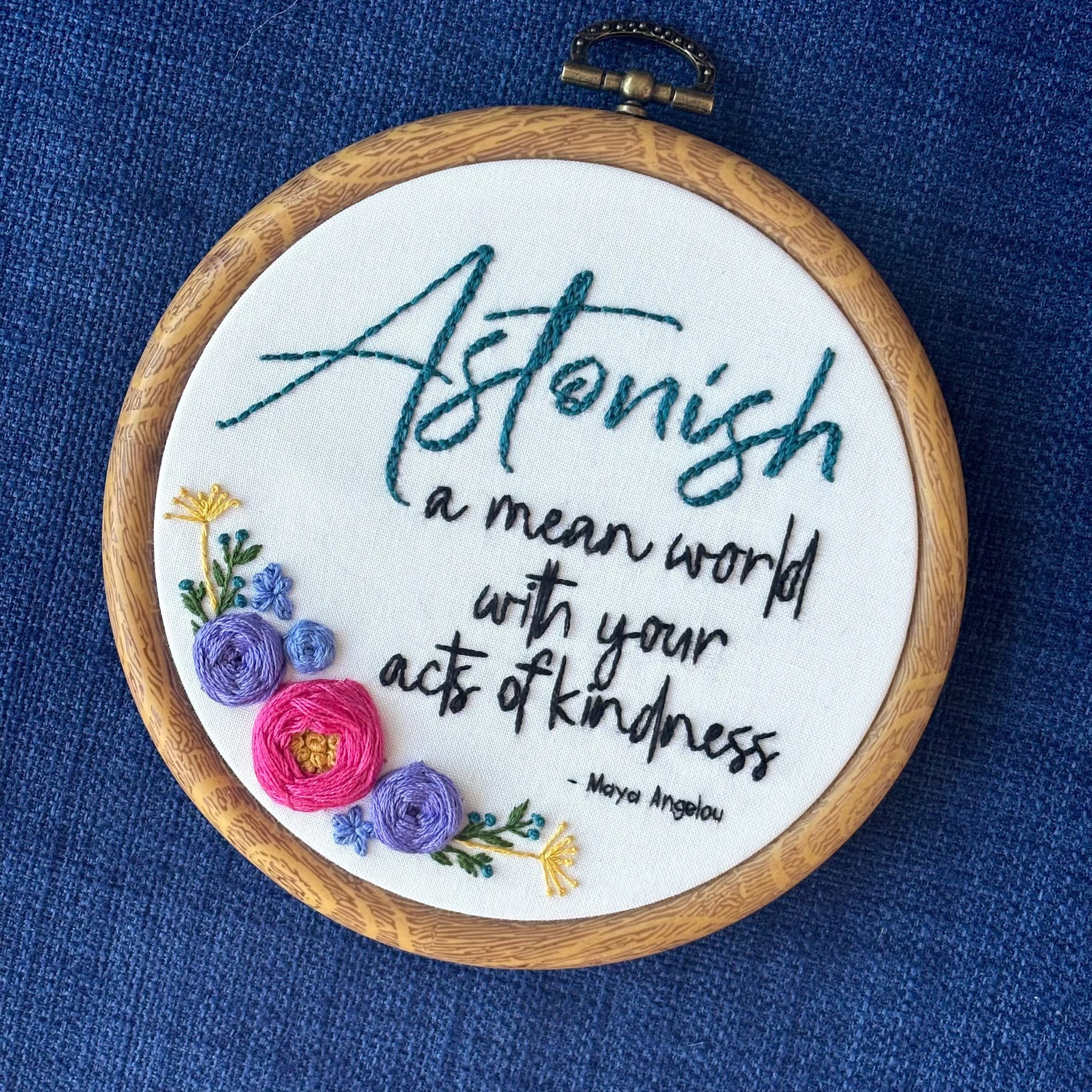 "Astonish a mean world with your acts of kindness. Maya Angelou." Astonish hand embroidered in dark teal. Remaining quote and attribution hand embroidered in black. In the lower left side, a cluster of flowers with bright pink and purple roses, golden pointy flowers, and green stems with leaves and dark teal flower buds. Art is displayed in a medium dark grained wood-look embroidery hoop.