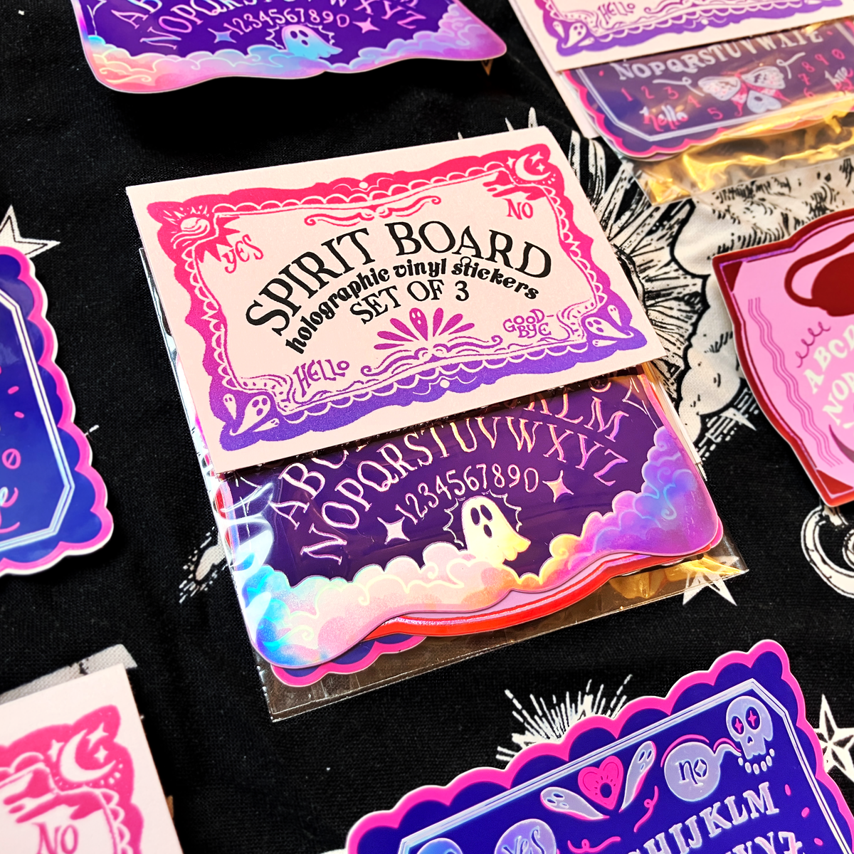 A set of three holographic vinyl stickers featuring gothic designs, including a Spirit Board, an Alphabet Board, and a Numbers Board, displayed on a black background with other stickers.