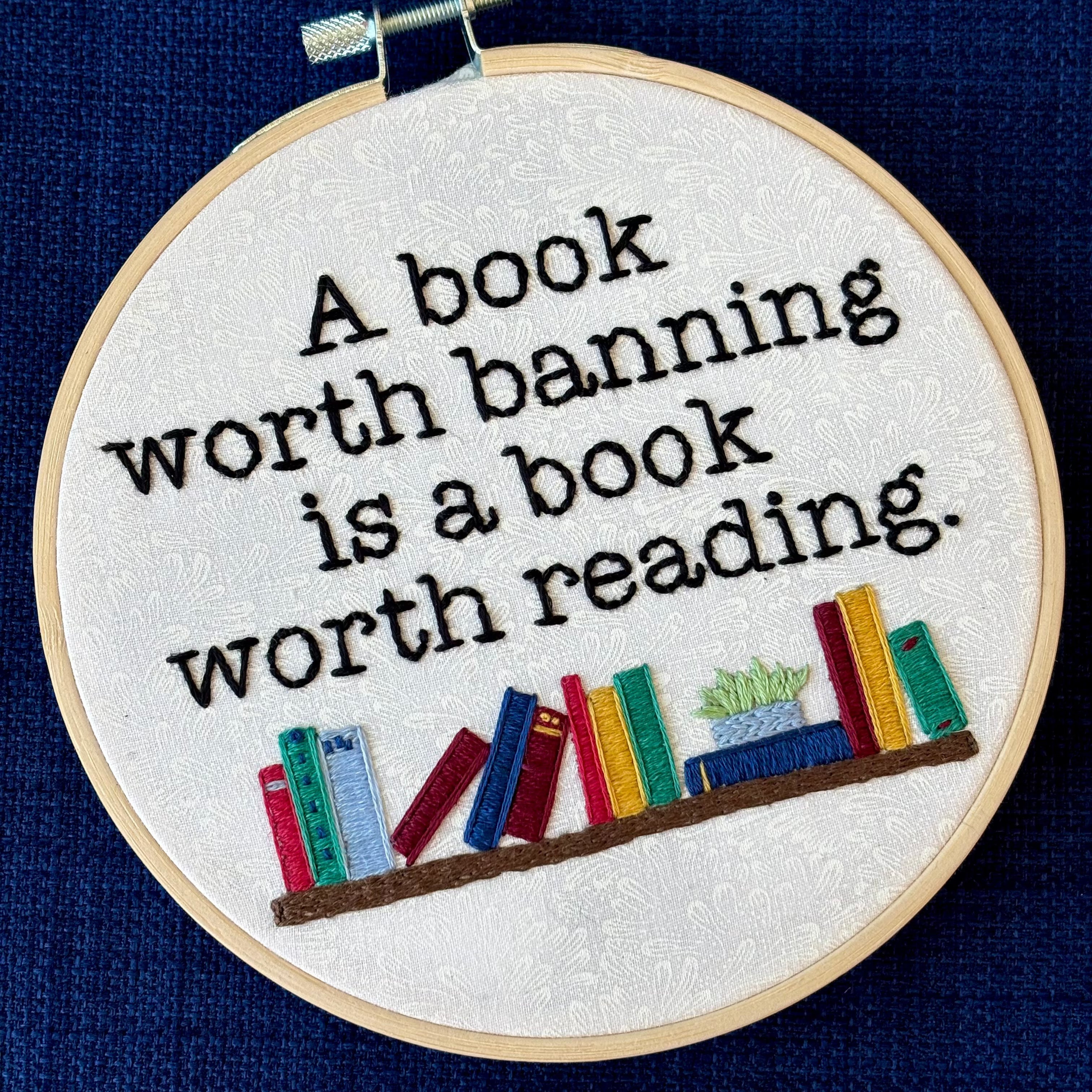 "A book worth banning is a book worth reading". Quote hand embroidered in black serif font. Below the text is an embroidered bookshelf with several different colored books, as well as a light blue planter holding a light green plant. Sewn on white-on-white cotton. Framed in a light wood embroidery hoop as wall art or bookshelf art.