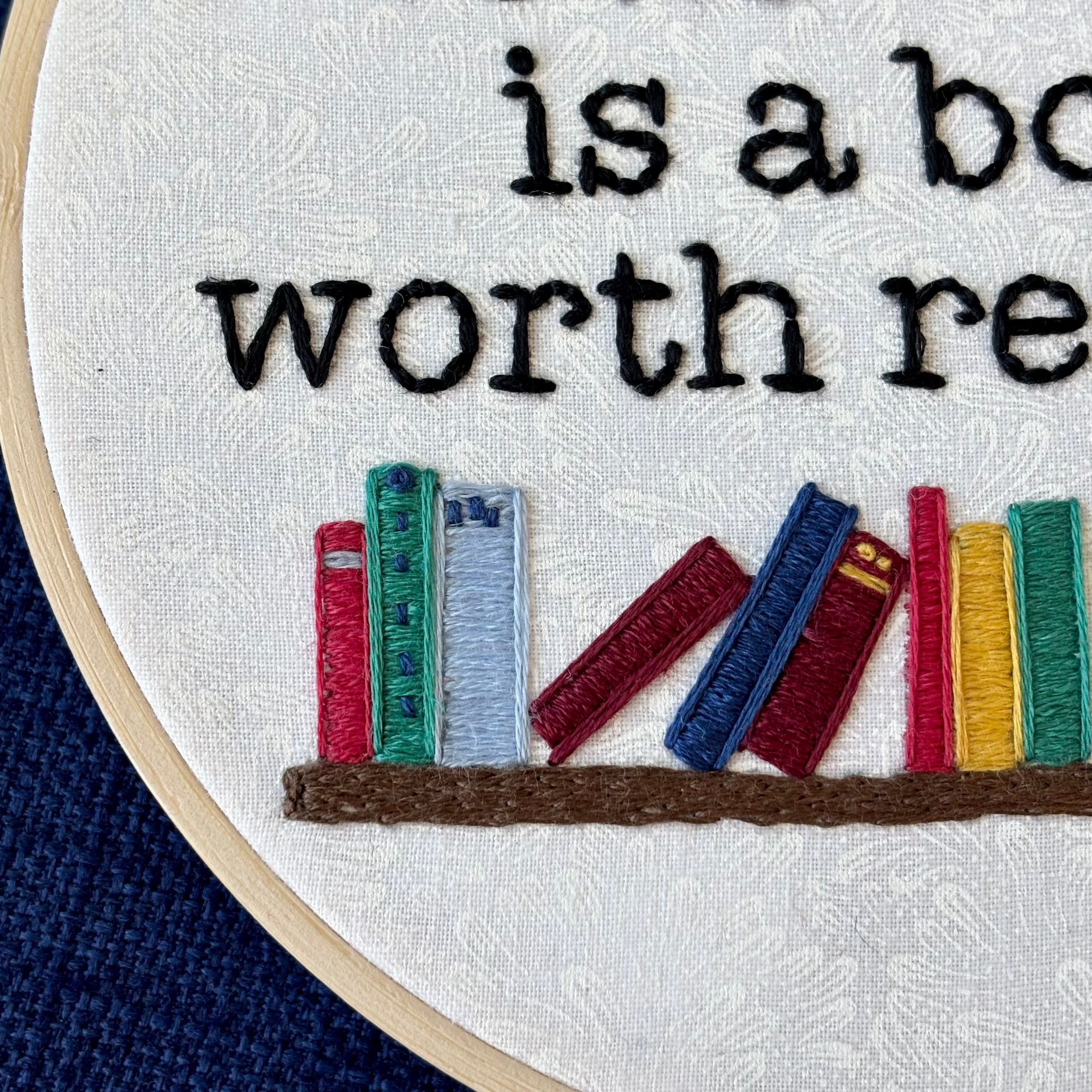 "A book worth banning is a book worth reading". Quote hand embroidered in black serif font. Below the text is an embroidered bookshelf with several different colored books, as well as a light blue planter holding a light green plant. Sewn on white-on-white cotton. Framed in a light wood embroidery hoop as wall art or bookshelf art.