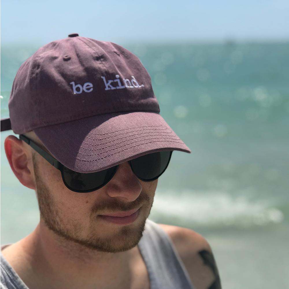 Purple cotton dad hat with white "be kind." embroidery worn outdoors by the water on a sunny day