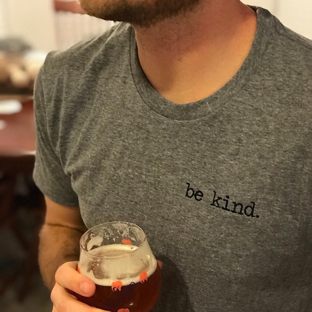 Heather gray short sleeve tee with black "be kind." embroidery on left chest, worn casually by a man holding a glass
