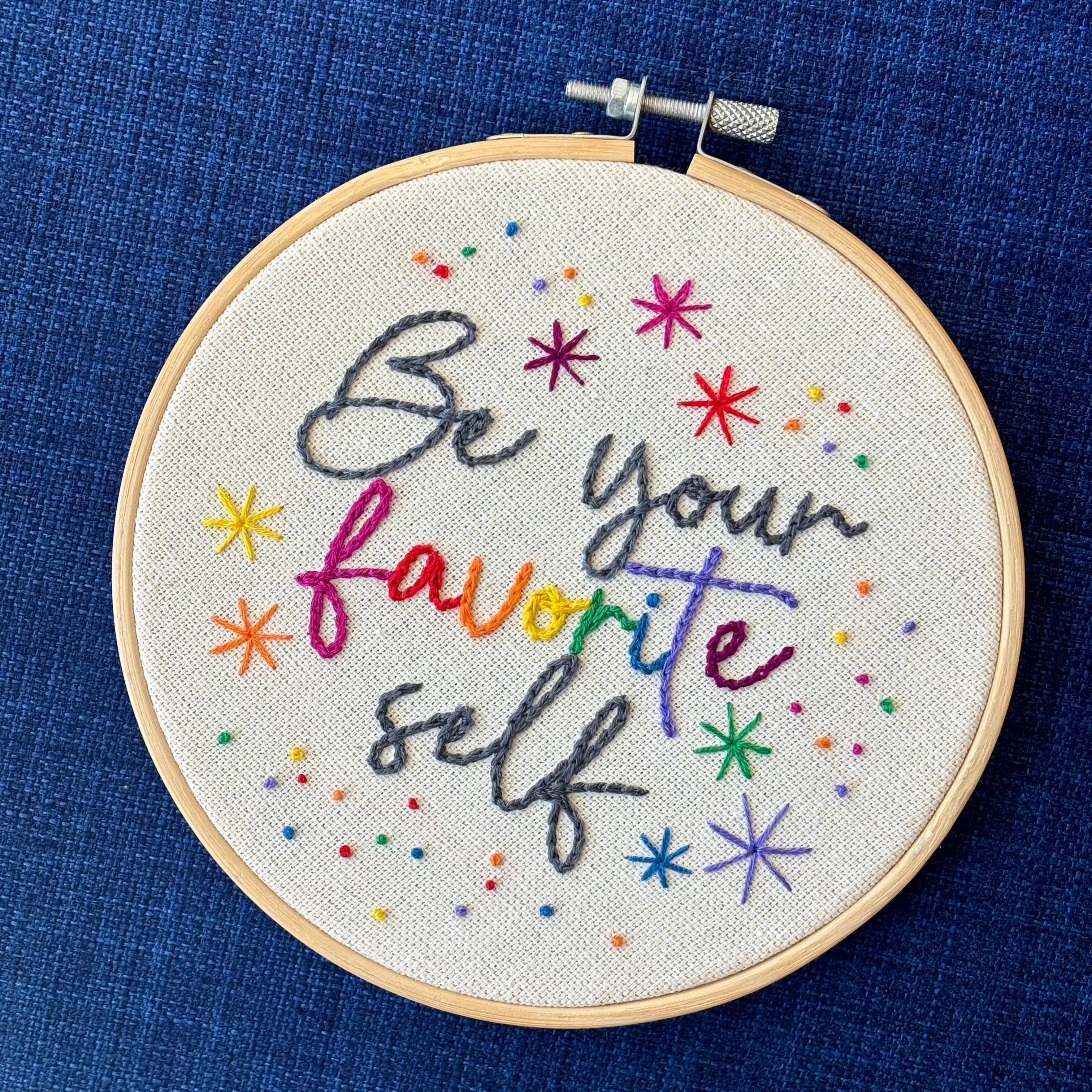 "Be Your Favorite Self" is hand embroidered in a script font on cream fabric. Favorite is sewn in rainbow colors, be your and self are embroidered in dark gray, and stars surround the quote. This wall art is framed in a light wood embroidery frame.
