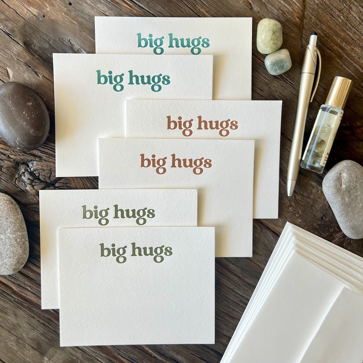 Staggered pile of six flat notecards, white in color with the words "big hugs" letterpress printed with embossed texture, two cards in teal ink, two in a warm orange and two in olive green. Styled on a wooden table with river rocks, a gold pen and a bottle of essential oil.
