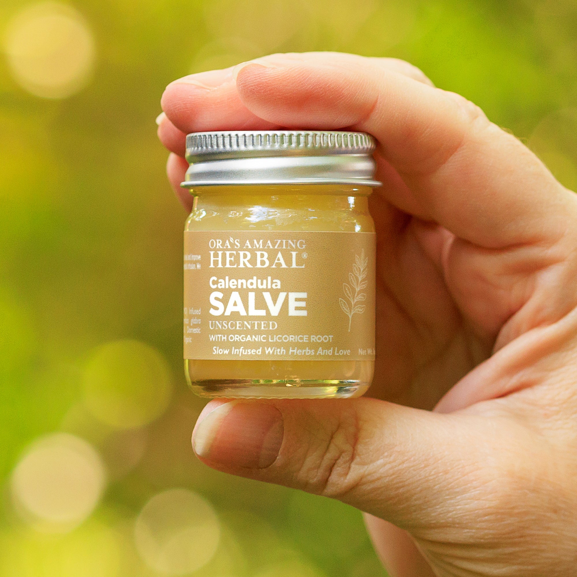 A hand holding a small glass jar of Ob’s Amazing Herbal Calendula Salve against a soft green bokeh background.