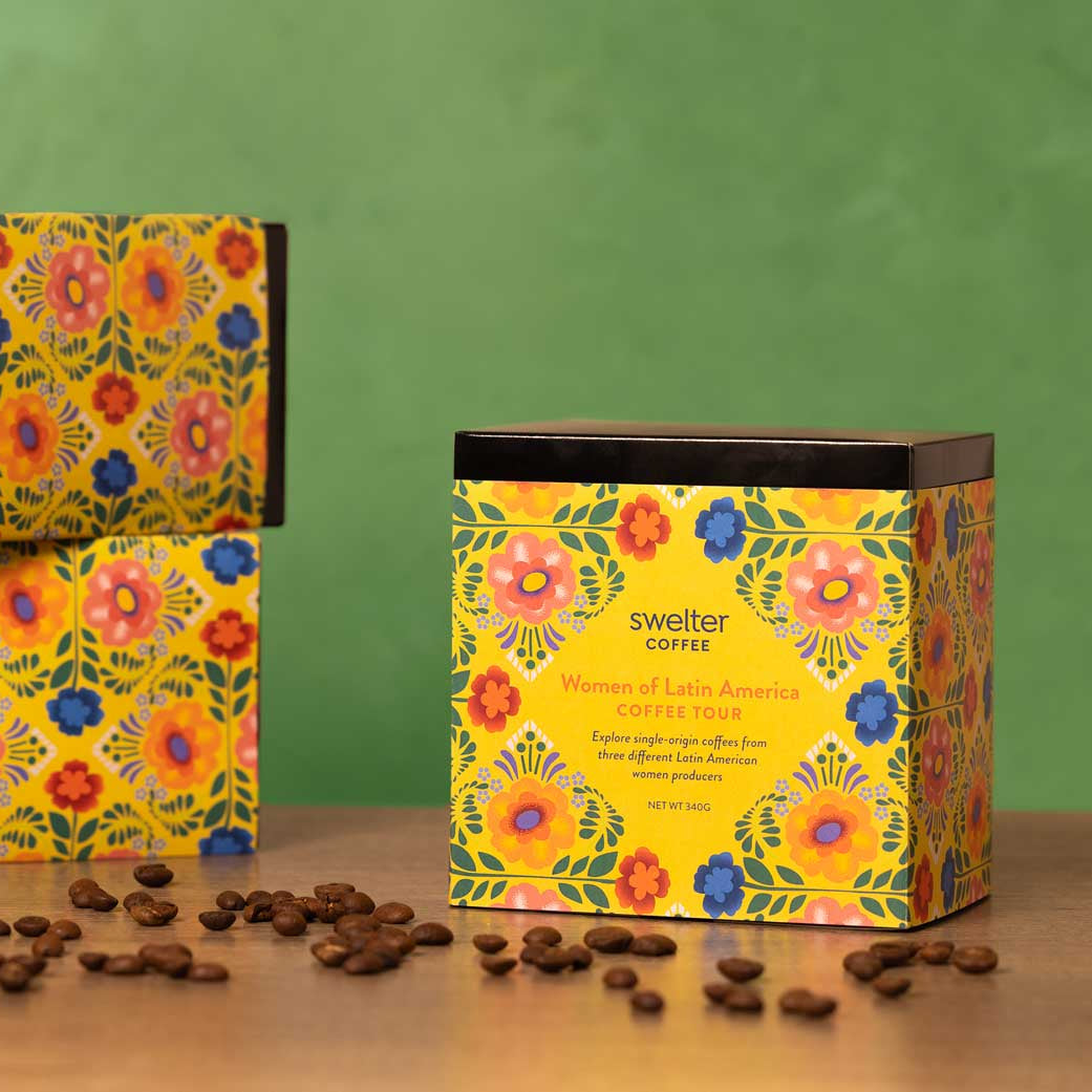 Three boxes of Swelter Coffee with floral designs on a wooden surface with scattered coffee beans, featuring women produced coffee from Latin American countries