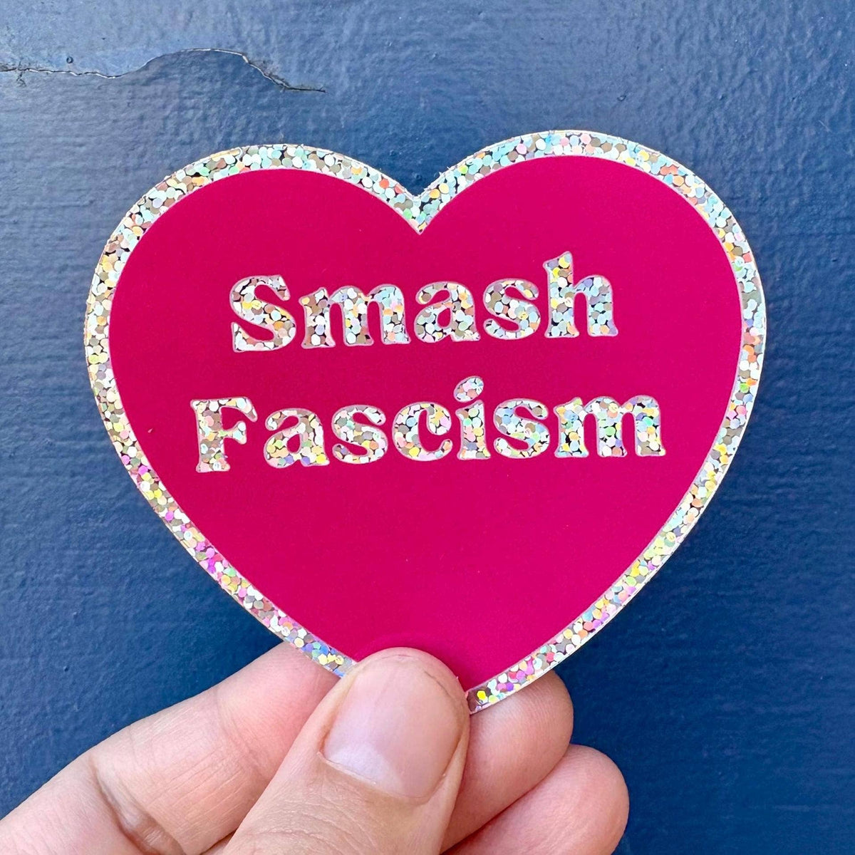 A hand holds a heart-shaped sticker with a bright pink background and a glittery multicolor border. The sticker features the text &