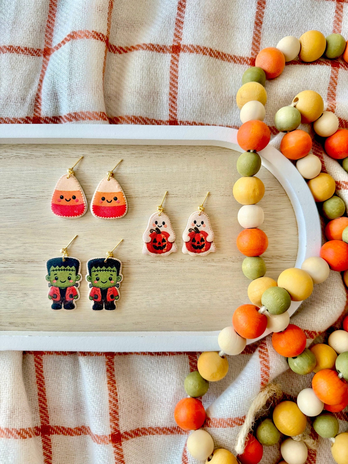 A styled flat-lay of Halloween-themed earrings displayed on a light wood tray, surrounded by a beaded garland on a cream plaid fabric background. The earrings include candy corn, jack-o&