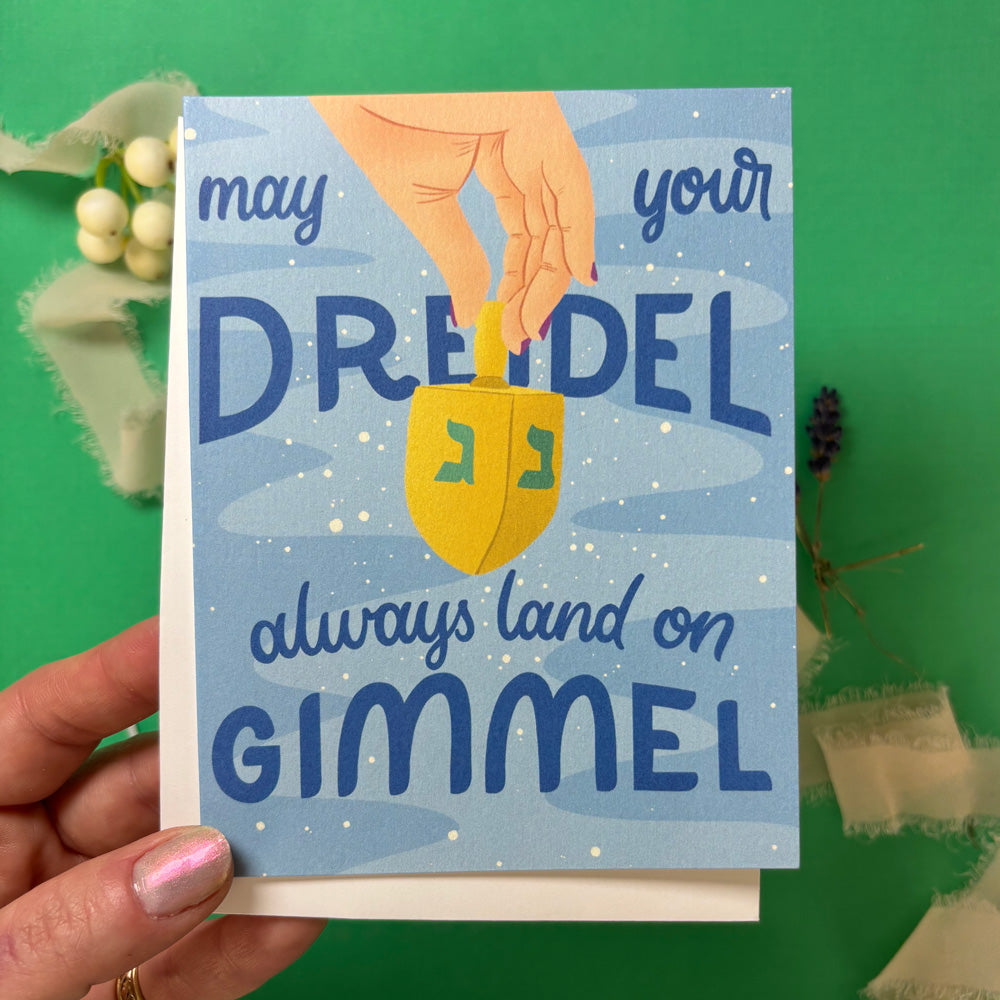 hand holding greeting Card with a dreidel illustration and text "may your dreidel always land on gimmel" on a green background with decorative elements.