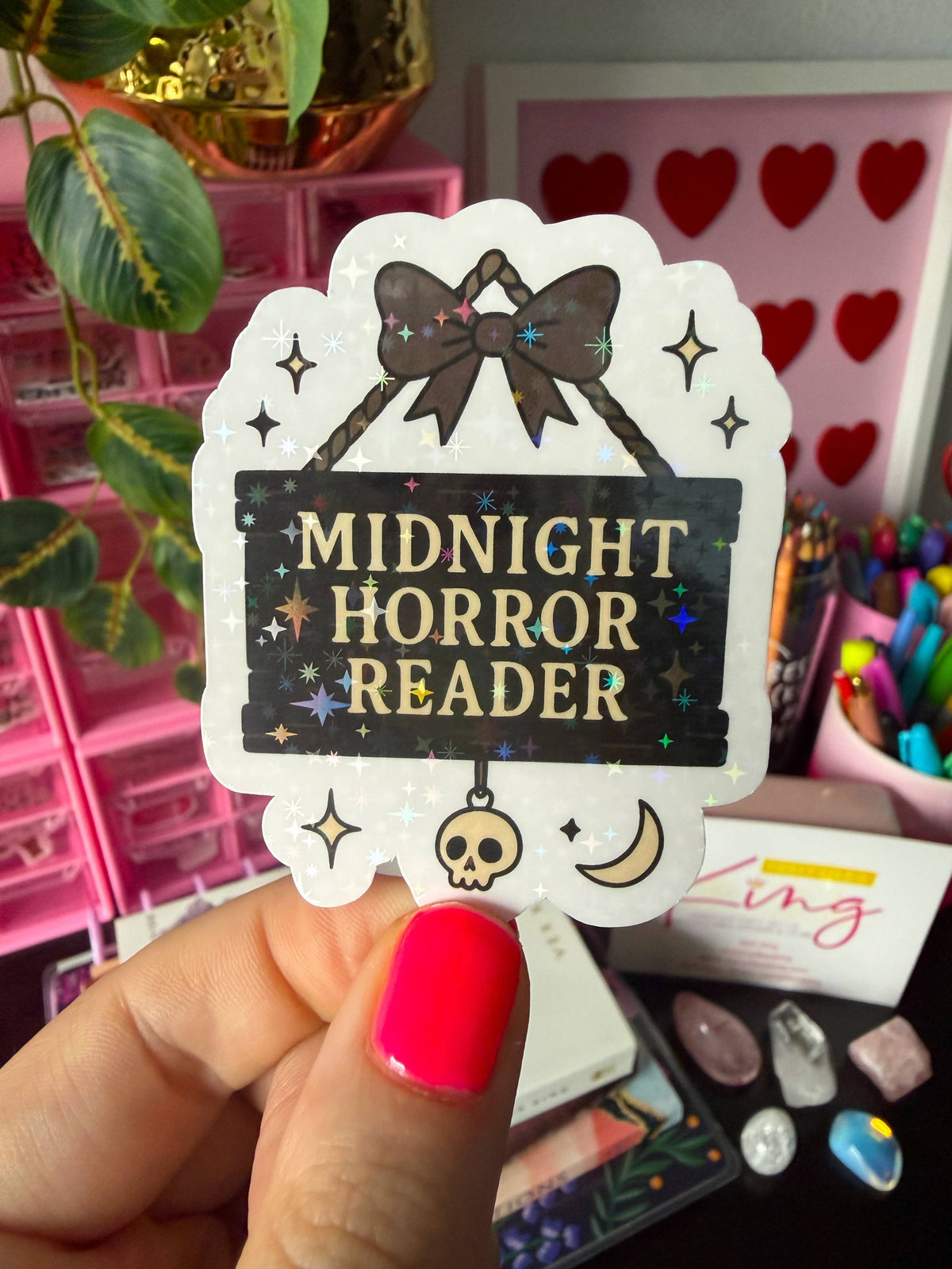 A hand with bright pink nail polish holds a die-cut sticker that reads &