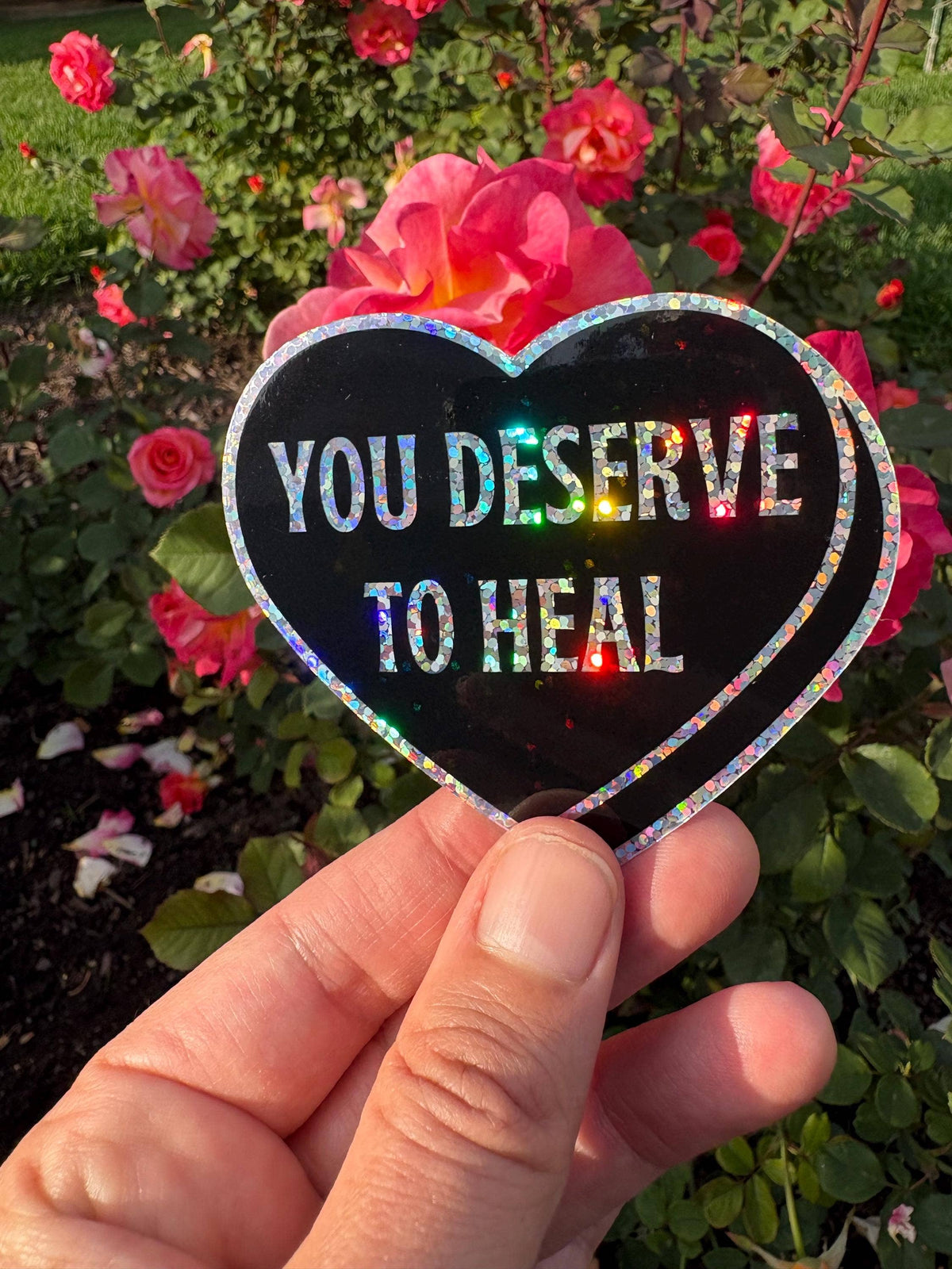 A hand holds a heart-shaped black sticker with holographic glitter edging and the text &