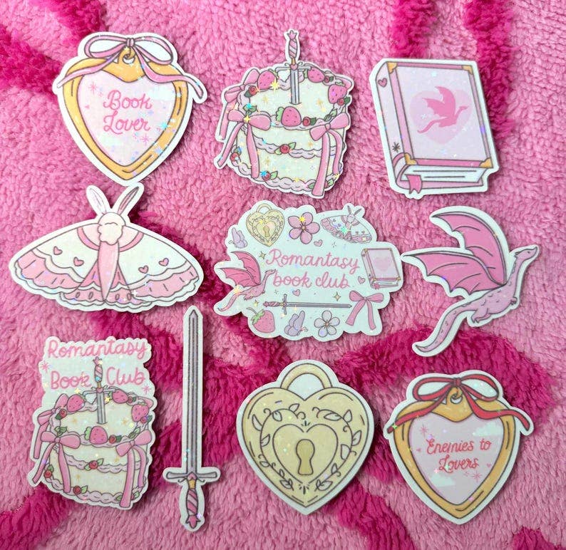 A set of cute, book-and-moth-themed enamel pins arranged on a pink textured fleece background. The pins feature illustrations of pink and cream-colored moths, a sword, a keyhole, and hearts with text such as &