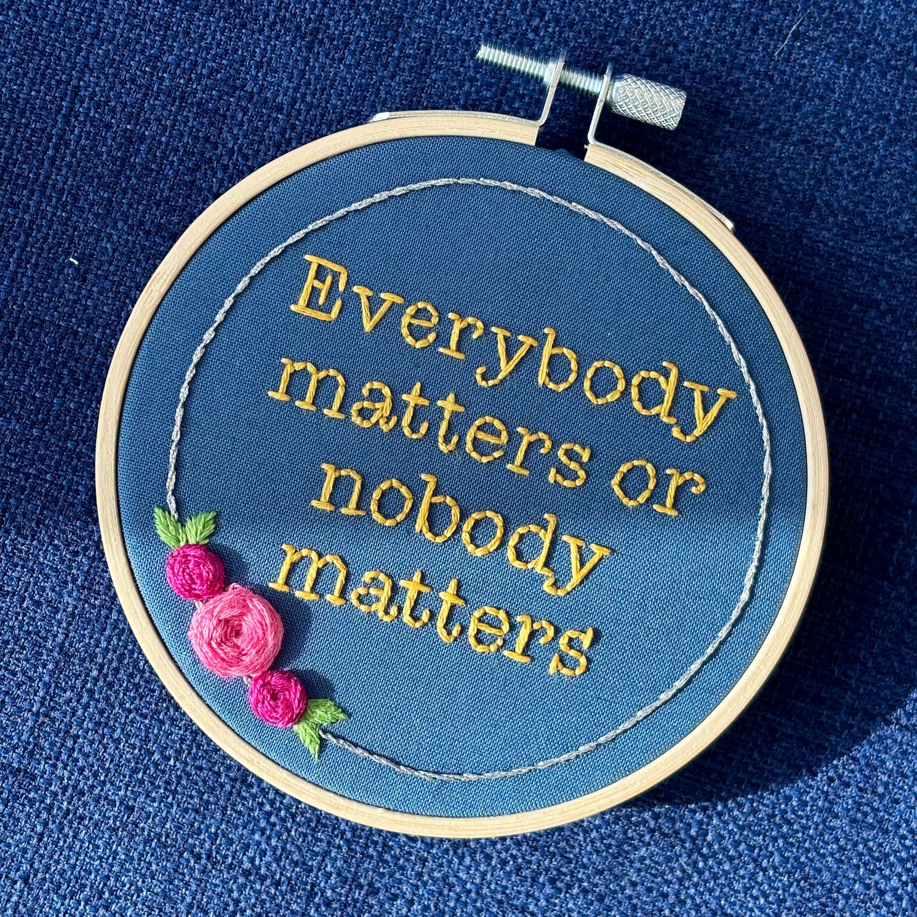"Everybody matters or nobody matters." Quote hand embroidered in sunny yellow thread in a serif typewriter font. Framed with a ring of metallic silver thread, spring green leaves, and pink and fuchsia roses. Stitched on blue cotton fabric and displayed in a light wooden embroidery hoop as wall art.