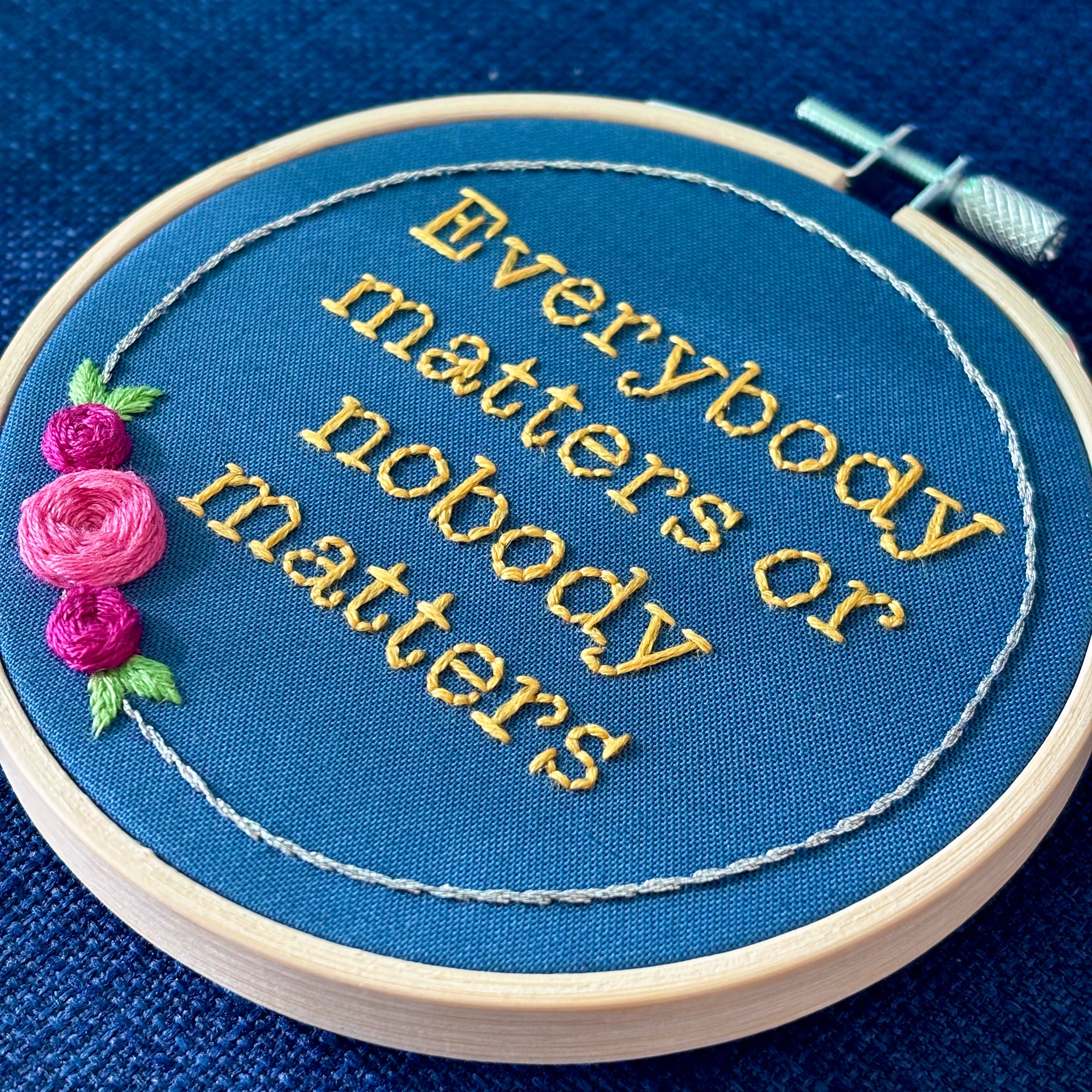 "Everybody matters or nobody matters." Quote hand embroidered in sunny yellow thread in a serif typewriter font. Framed with a ring of metallic silver thread, spring green leaves, and pink and fuchsia roses. Stitched on blue cotton fabric and displayed in a light wooden embroidery hoop as wall art.