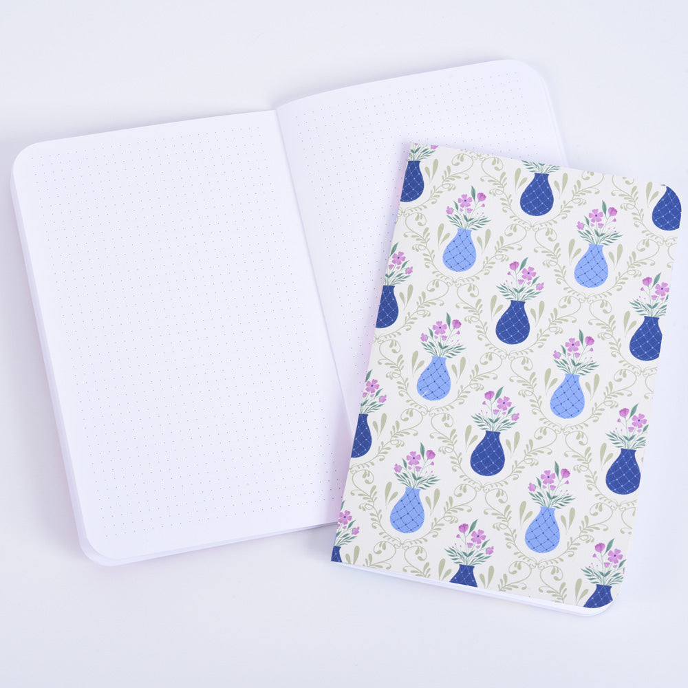 small pocket notebook with a pattern of blue vases and purple flowers on a sage green background, resting on an open notebook with dot grid pages