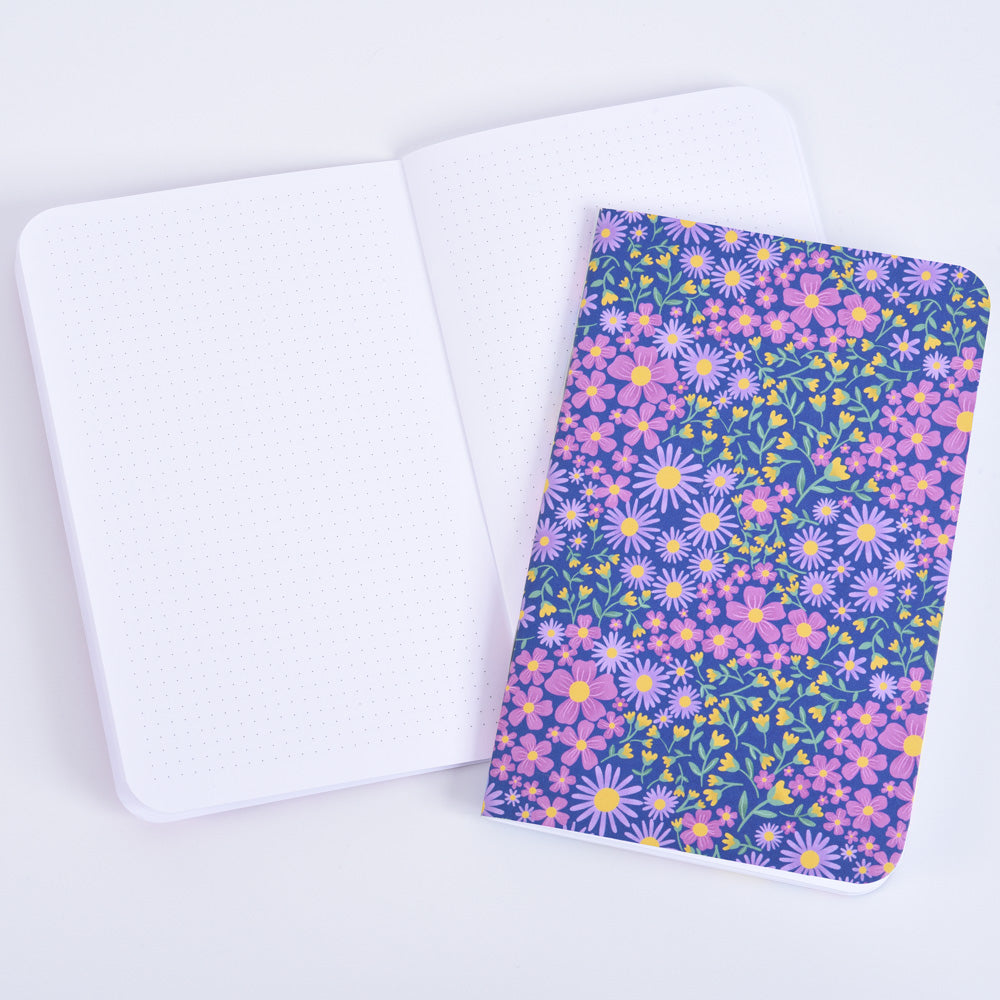 a closed small pocket notebook featuring a floral design on dark blue background, on top of an open notebook with small dot grid pages