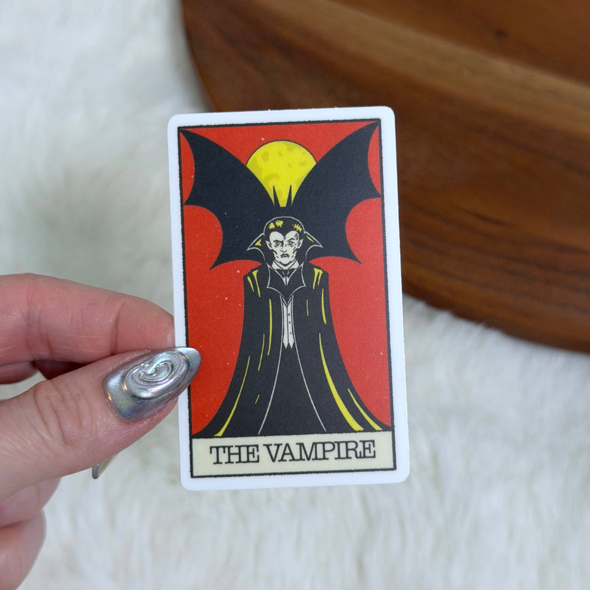 A hand with silver nail polish holds a tarot card featuring &