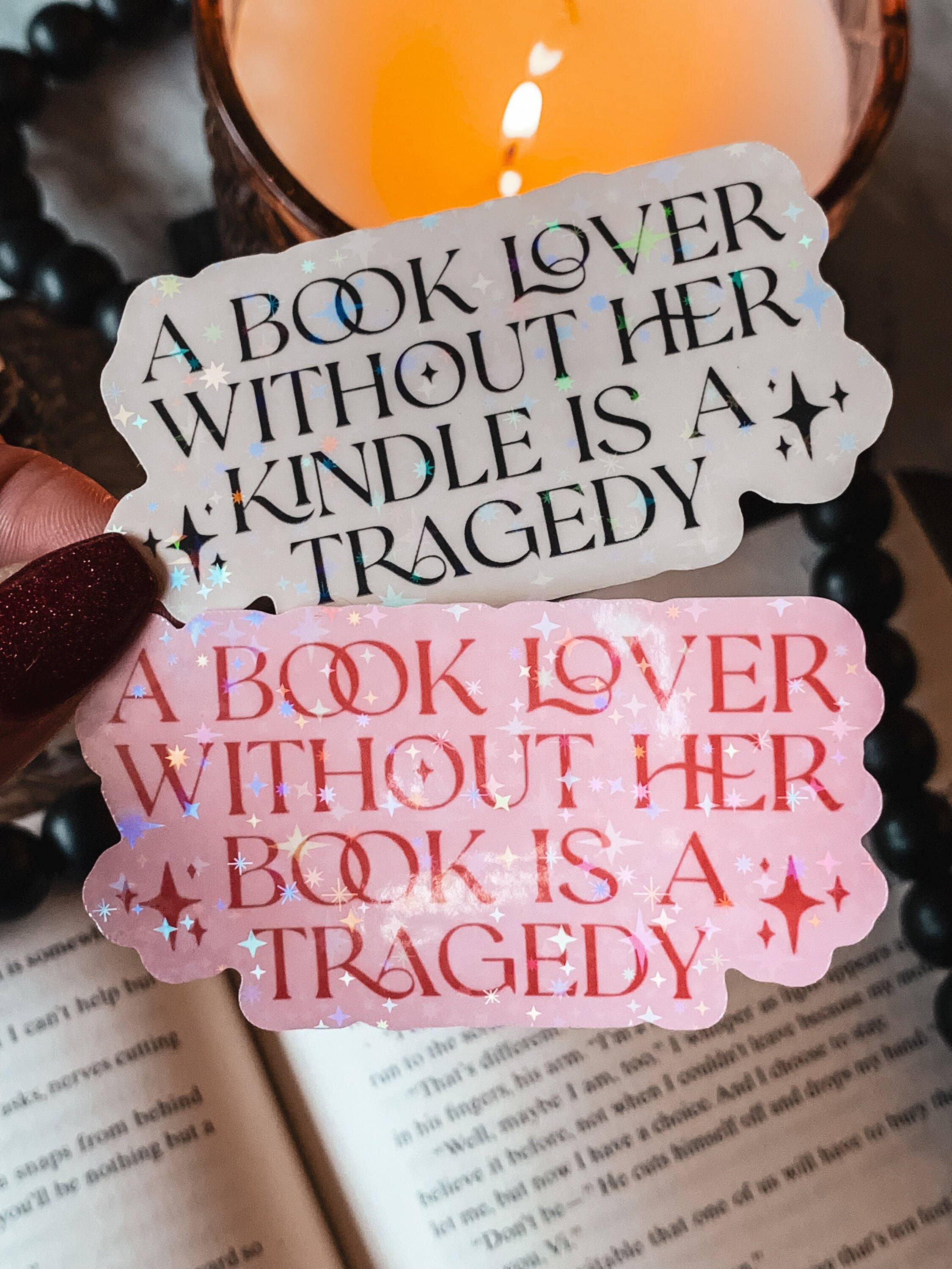 Two stickers with book-themed text held over an open book, with a candle in the background.