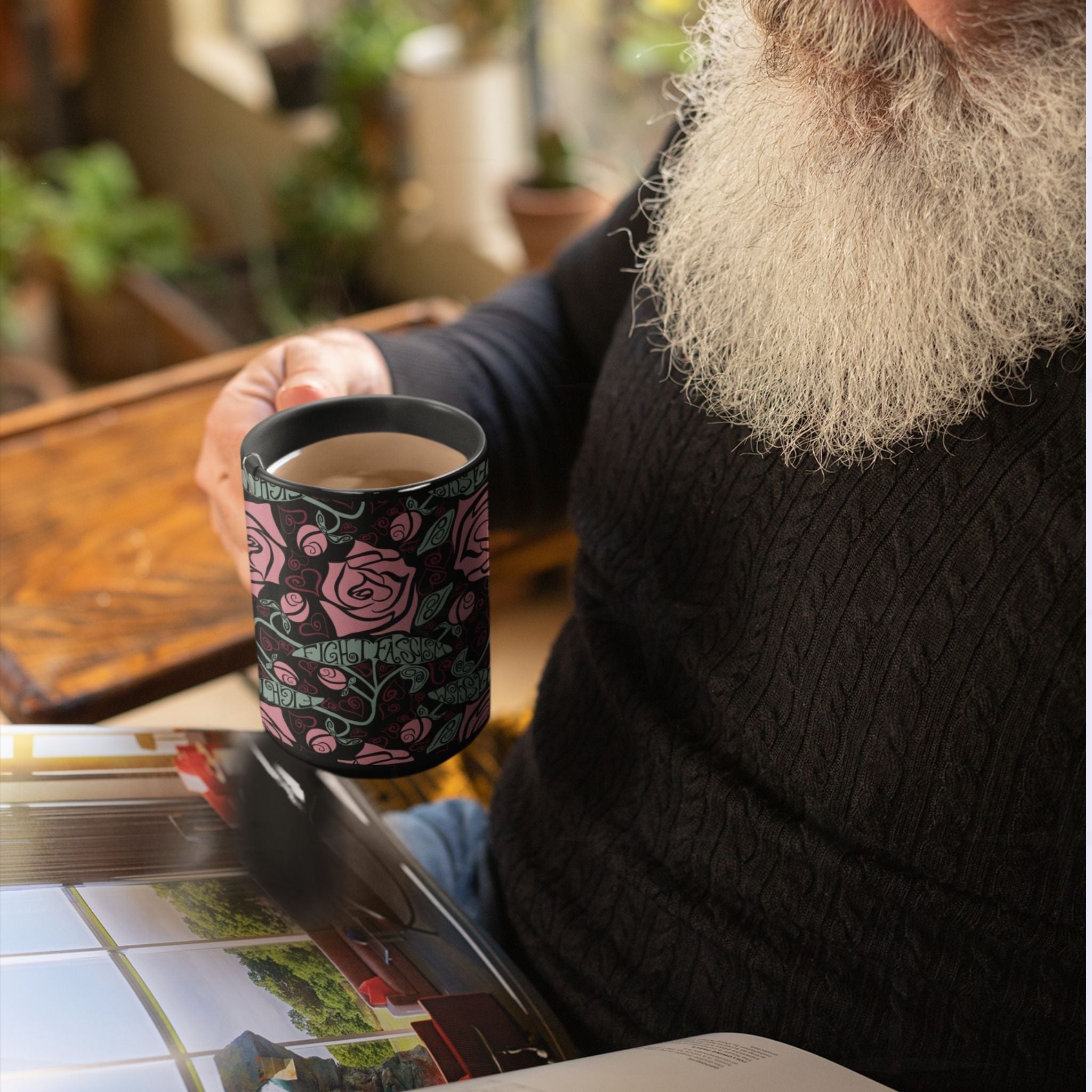 Person reading a magazine with the Fight Fascism black ceramic mug on a table, Art Nouveau floral wrap print visible