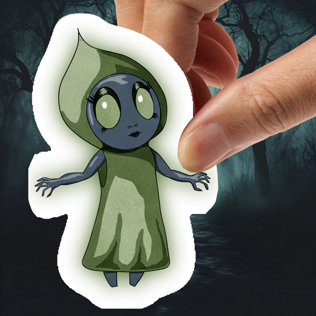 Hand holding the Flatwoods Monster sticker, a gray figure in a green hood with large pale eyes, against dark woods