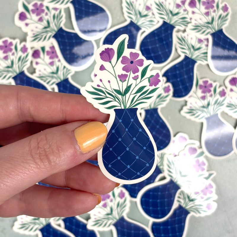 hand holding a sticker of a blue flower vase with purple flowers