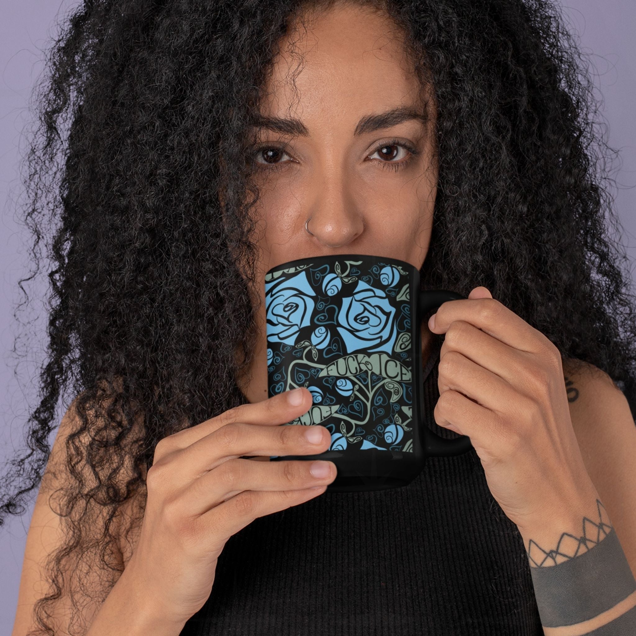 Person with curly hair holding a dark floral activist mug up to their face, showing the full wrap-around blue rose and Fuck ICE leaf pattern on glossy black ceramic