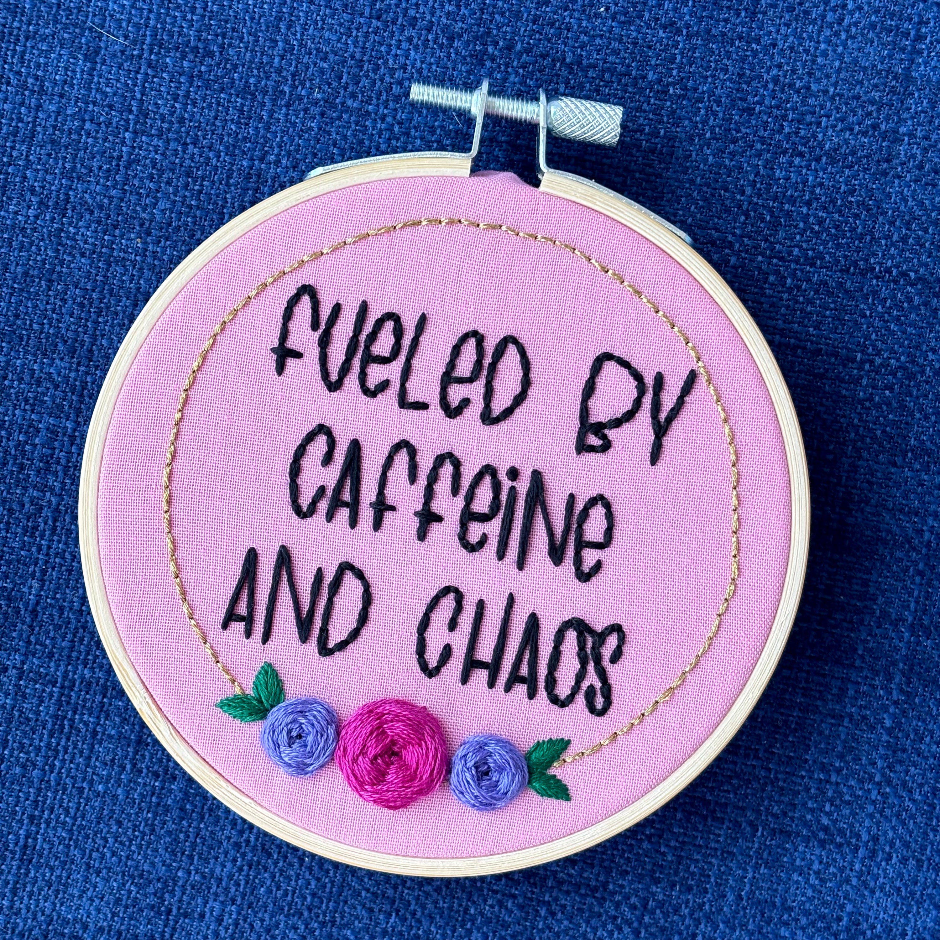 Finished embroidery hoop reads, "fueled by caffeine and chaos". Quote in handwriting print font and hand embroidered in black. Words are centered in a floral frame, which is metallic gold and embellished with cheerful pink and purple roses and dark green leaves sewn with embroidery thread. Fabric is pink cotton. Framed in light wood embroidery hoop as wall art.