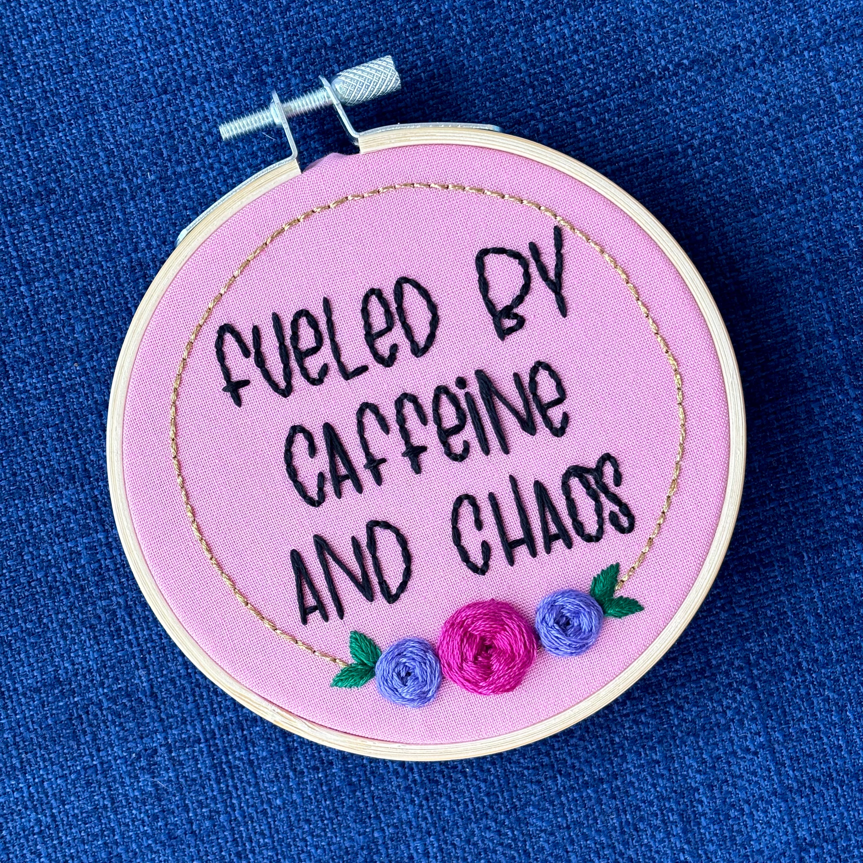 Finished embroidery hoop reads, "fueled by caffeine and chaos". Quote in handwriting print font and hand embroidered in black. Words are centered in a floral frame, which is metallic gold and embellished with cheerful pink and purple roses and dark green leaves sewn with embroidery thread. Fabric is pink cotton. Framed in light wood embroidery hoop as wall art.
