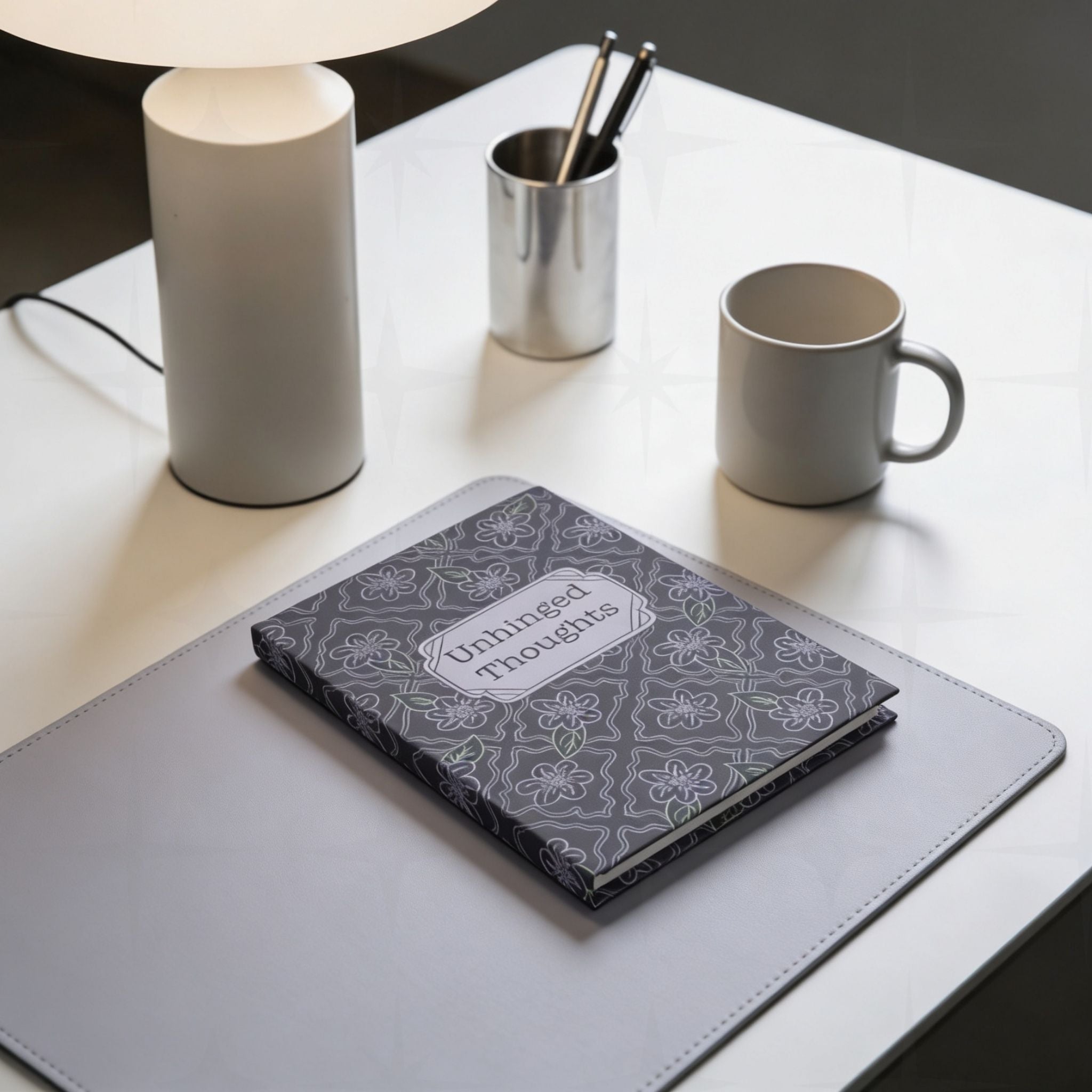 Unhinged Thoughts gothic floral journal on grey leather desk pad beside a table lamp and coffee mug, modern workspace setting