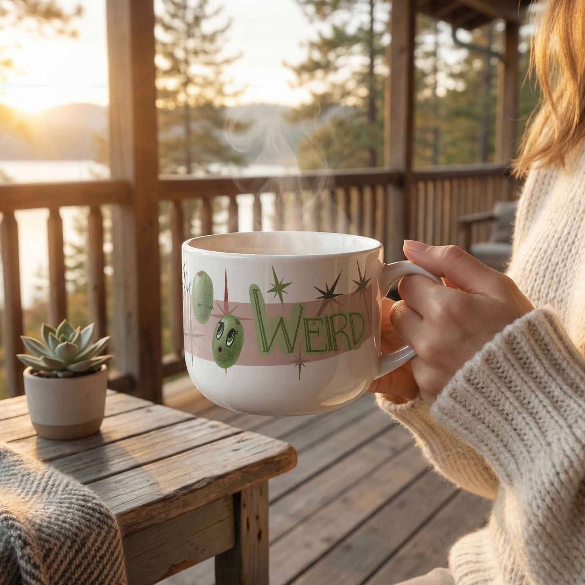 Woman in cream knit sweater holding 25oz funny mom mug with steam rising on a rustic cabin porch at golden hour, retro Olive My Kids Are Weird olive pun design facing forward