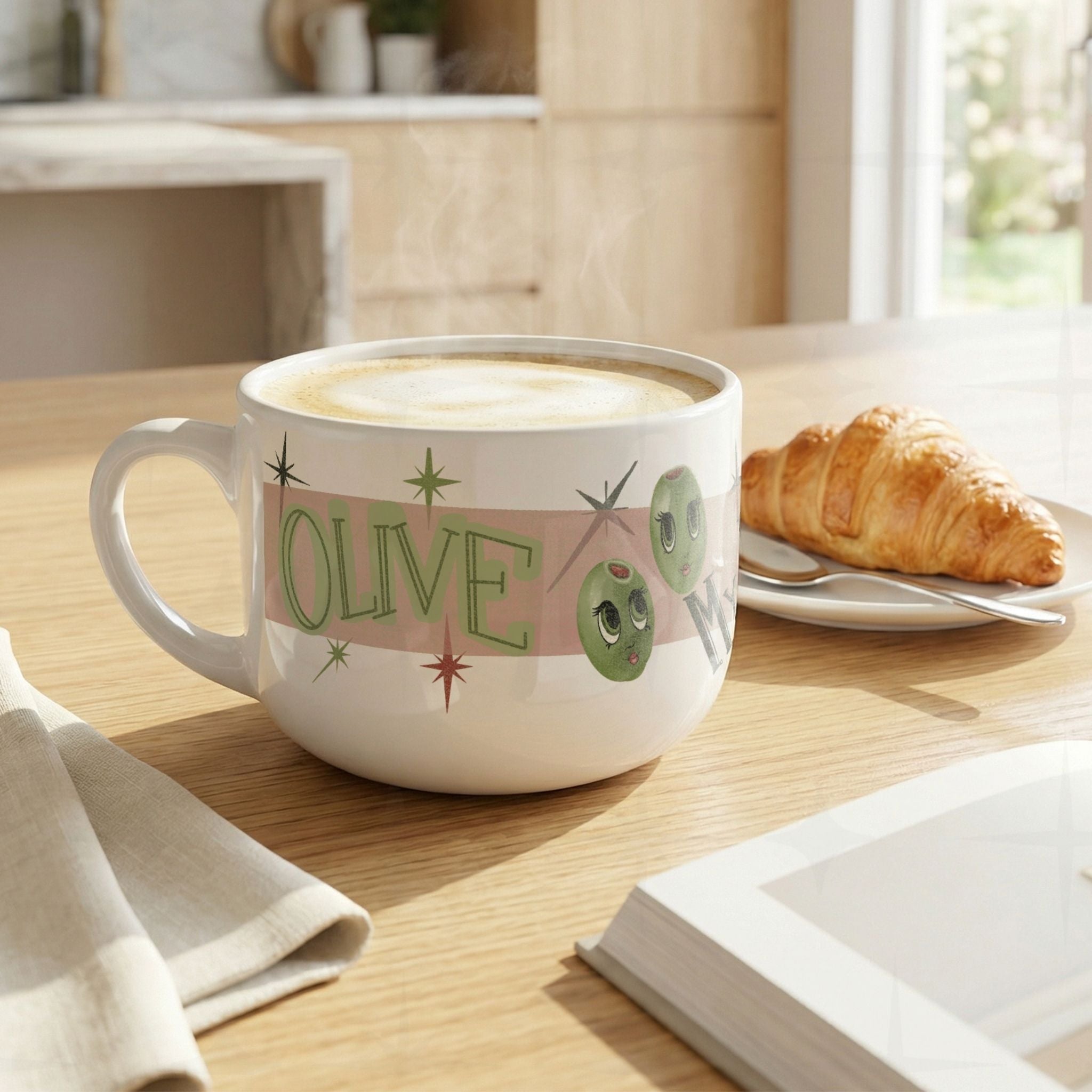 25oz oversized white ceramic funny mom mug with latte foam on wood kitchen table beside croissant, Olive My Kids Are Weird retro olive pun design with mid-century starburst details visible