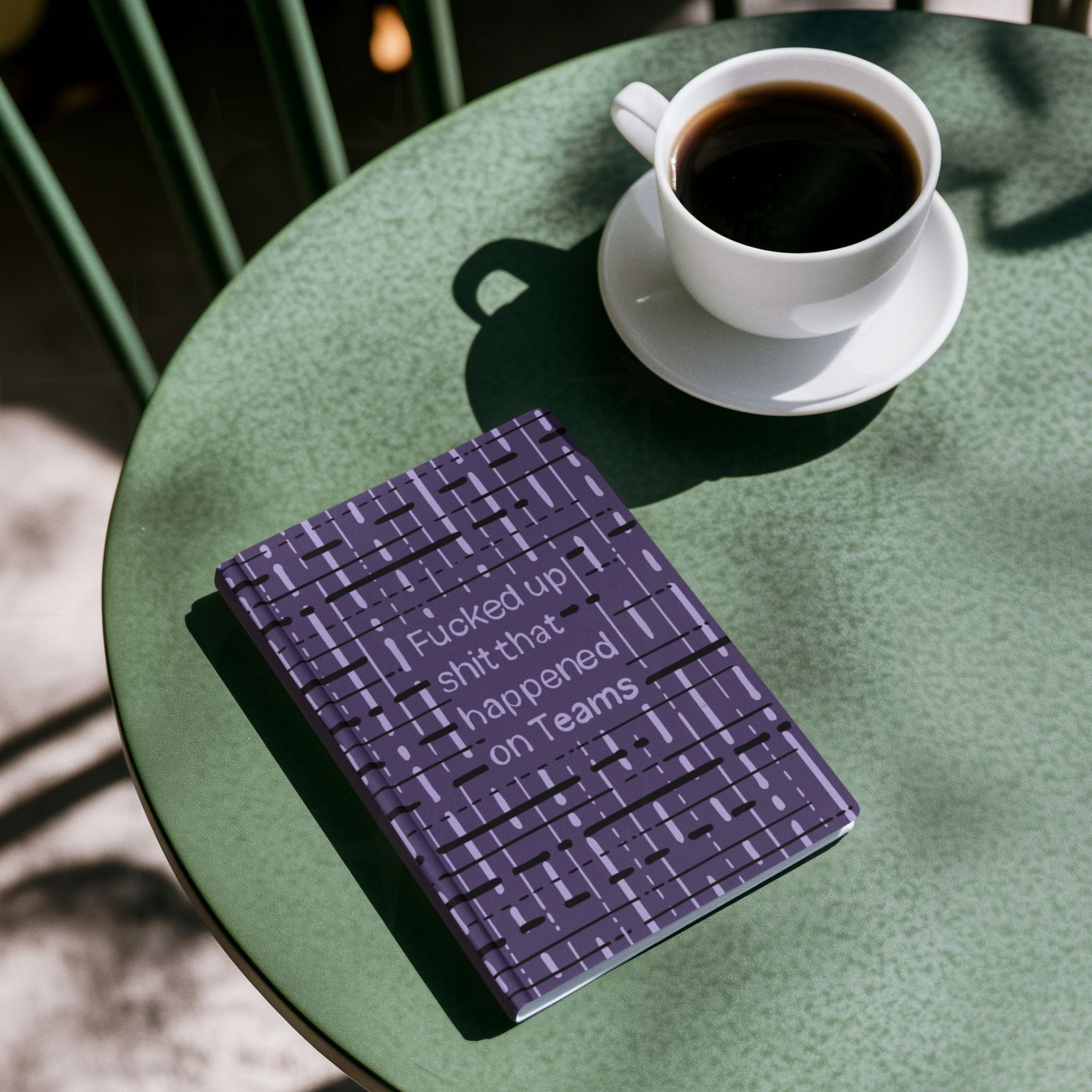 Funny Teams profanity hardcover journal on green café table next to cup of black coffee