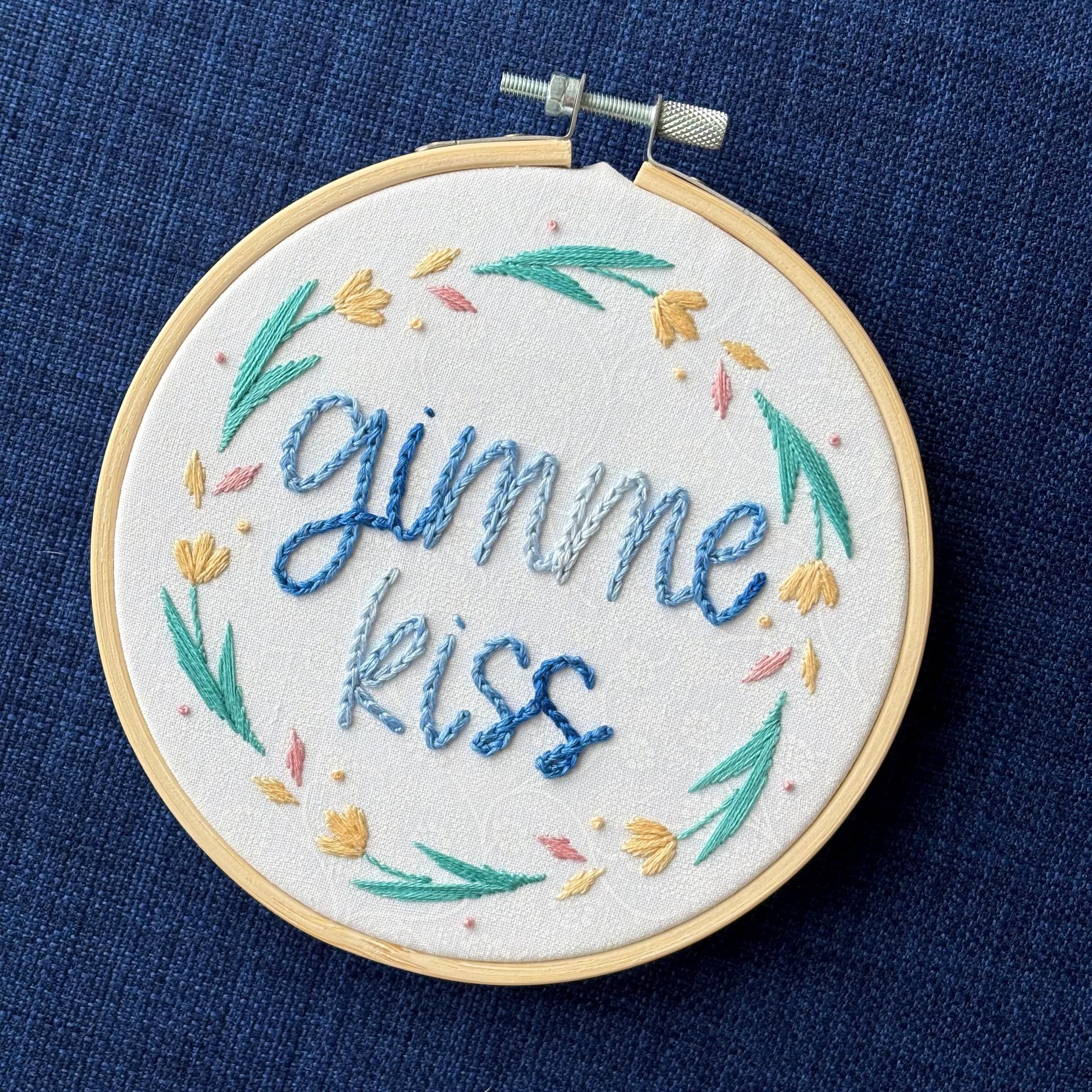 "Gimme kiss". Quote hand embroidered in handwriting font in variegated blues. Surrounding the text is a floral wreath with pastel yellow flowers, green stems and leaves, and yellow and pink floating petals. Sewn on white-on-white cotton. Framed as wall art in a light wood embroidery hoop.
