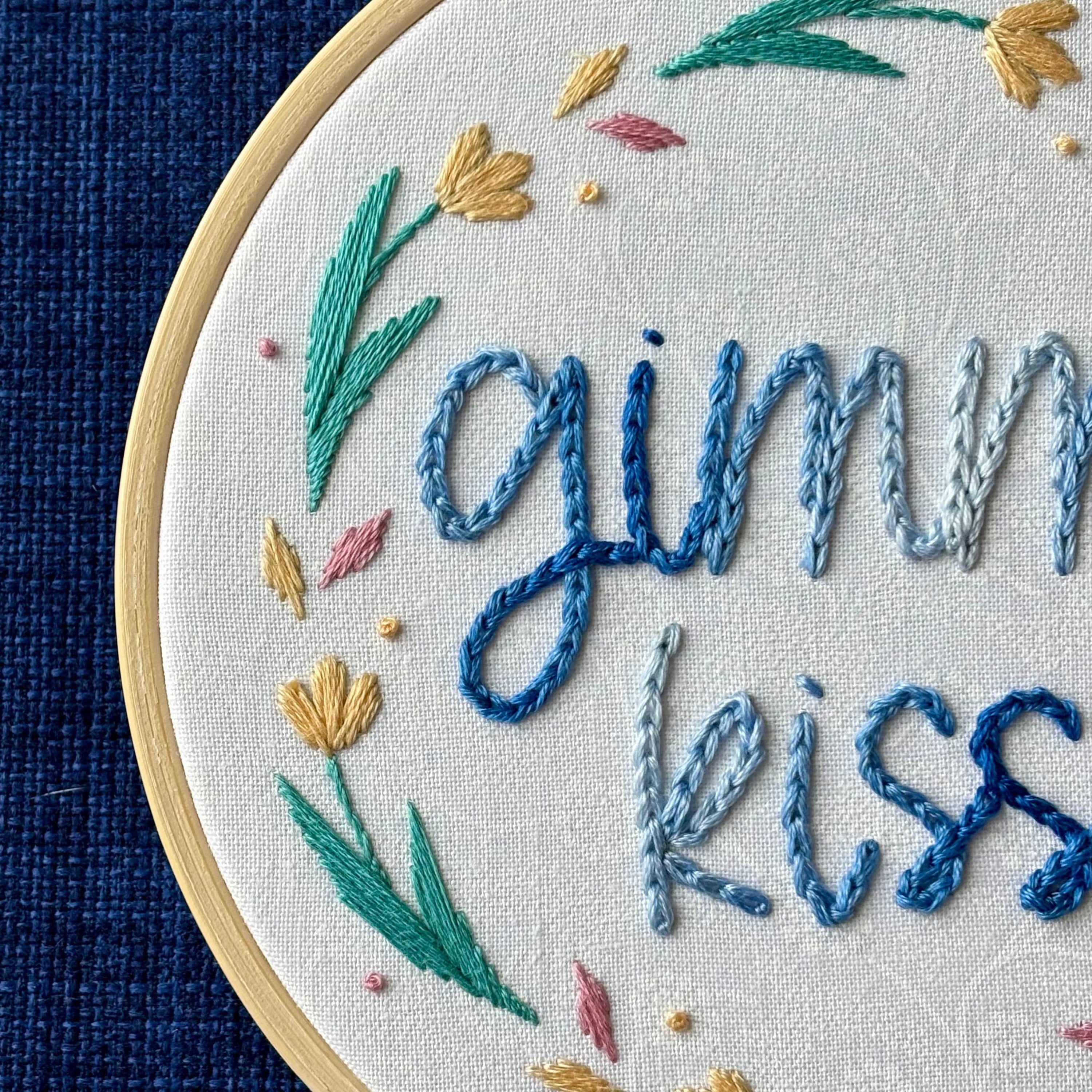"Gimme kiss". Quote hand embroidered in handwriting font in variegated blues. Surrounding the text is a floral wreath with pastel yellow flowers, green stems and leaves, and yellow and pink floating petals. Sewn on white-on-white cotton. Framed as wall art in a light wood embroidery hoop.