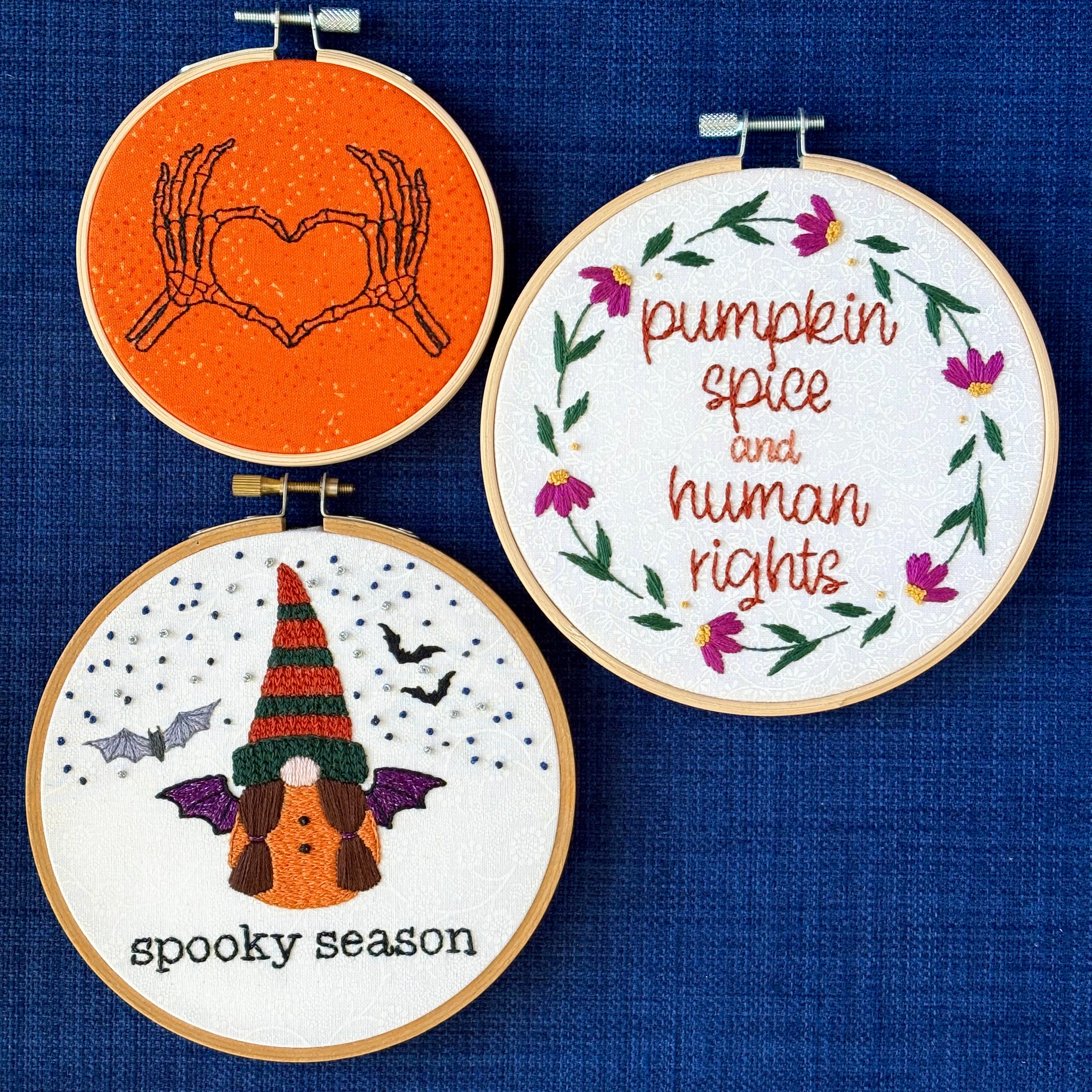 Hand embroidered pair of skeleton hands making a heart by touching thumbs and forefingers, while remaining fingers fan out. "Pumpkin spice and human rights". Handwriting font text surrounded by a floral wreath of pink asters and green leaves and stems. Hand embroidered gnome wears a an orange robe with black and sparkly purple bat wings. Under the gnome is "spooky season" embroidered in sparkling black thread. 3 designs displayed in light wooden hoops as wall art or Christmas ornaments.
