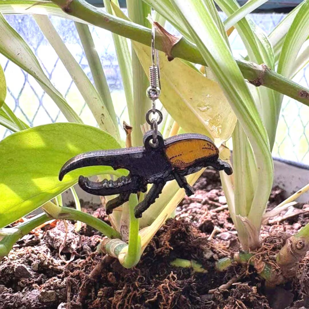 Close-up side view of hand-painted wooden hercules beetle dangle earrings showing layered wood construction, black body, golden wing covers, and hypoallergenic .925 sterling silver hooks.