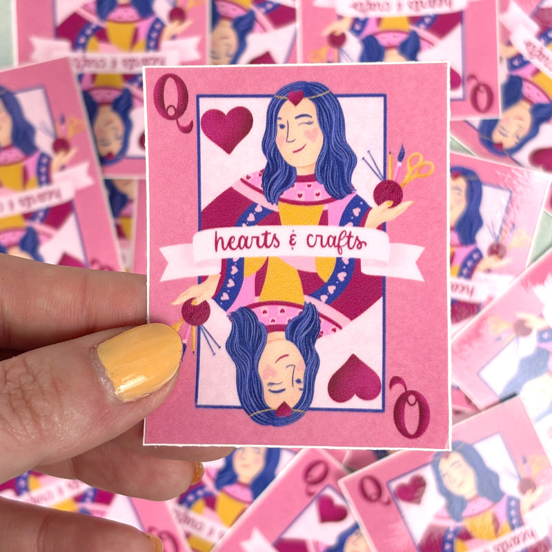 hand holding A pink sticker with a design of a queen playing card surrounded by hearts and crafting tools. The text &