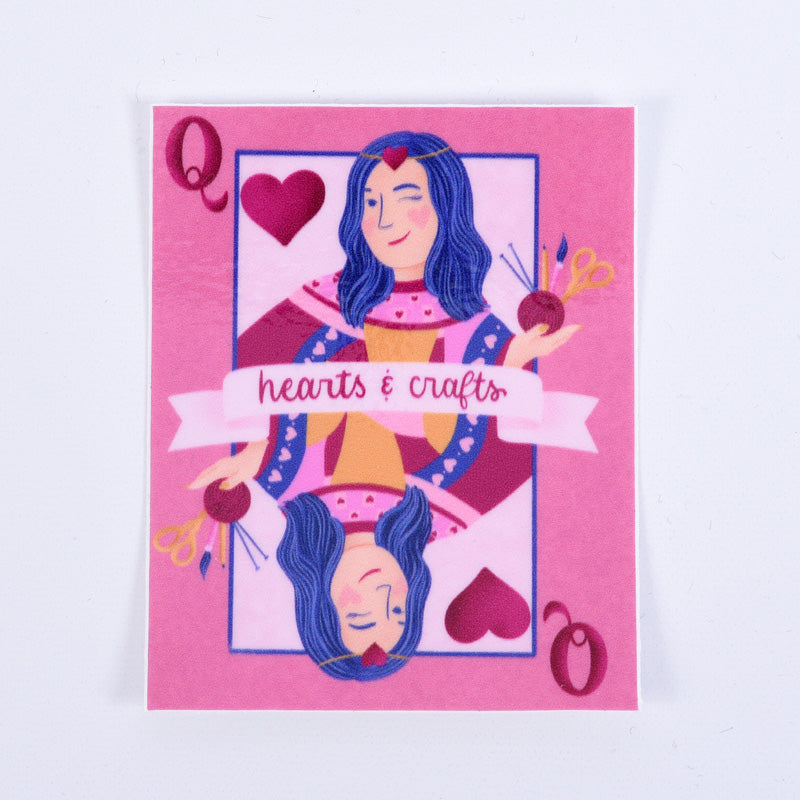 A pink sticker with a design of a queen playing card surrounded by hearts and crafting tools. The text &
