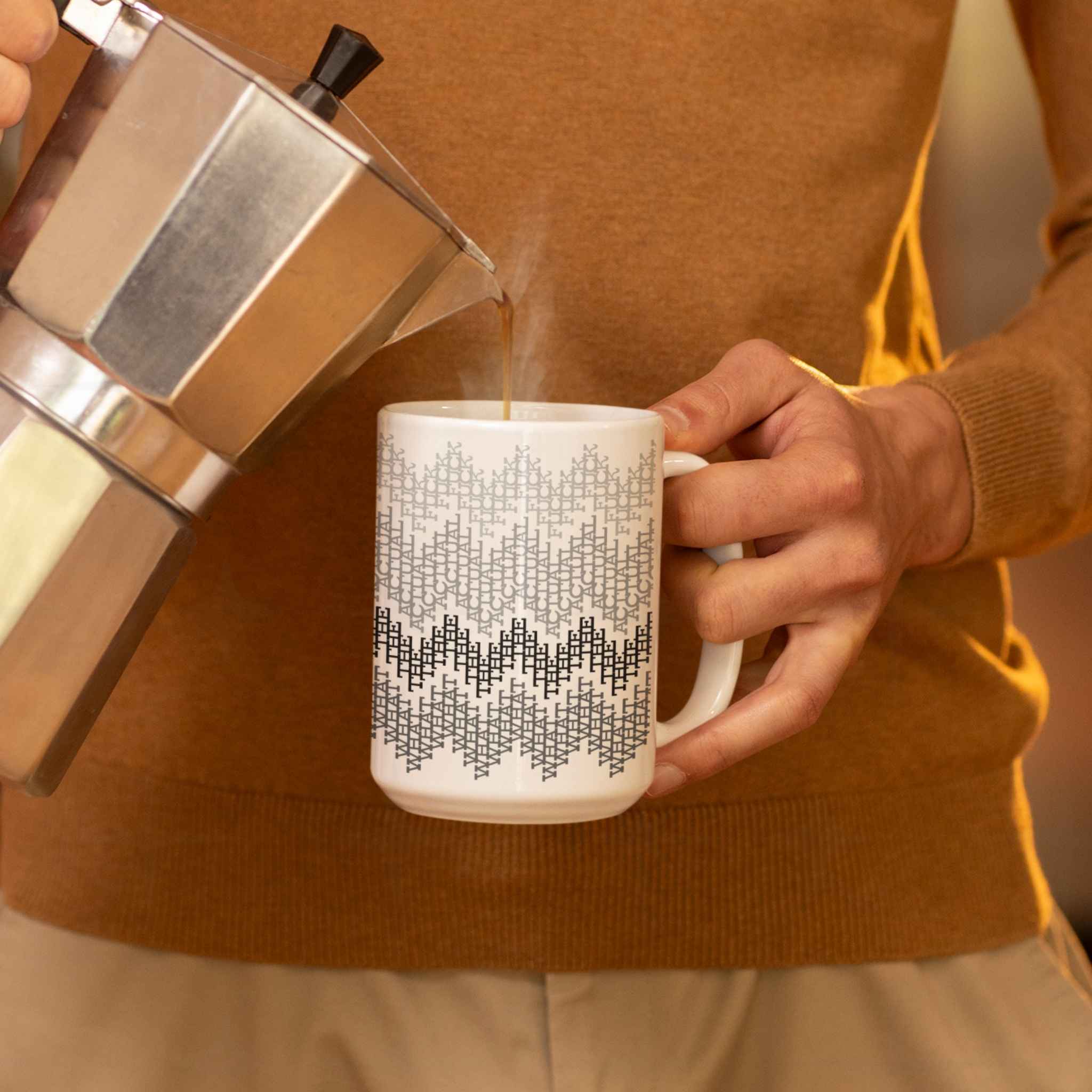 Coffee being poured from a moka pot into a gray hidden profanity mug, warm indoor morning setting