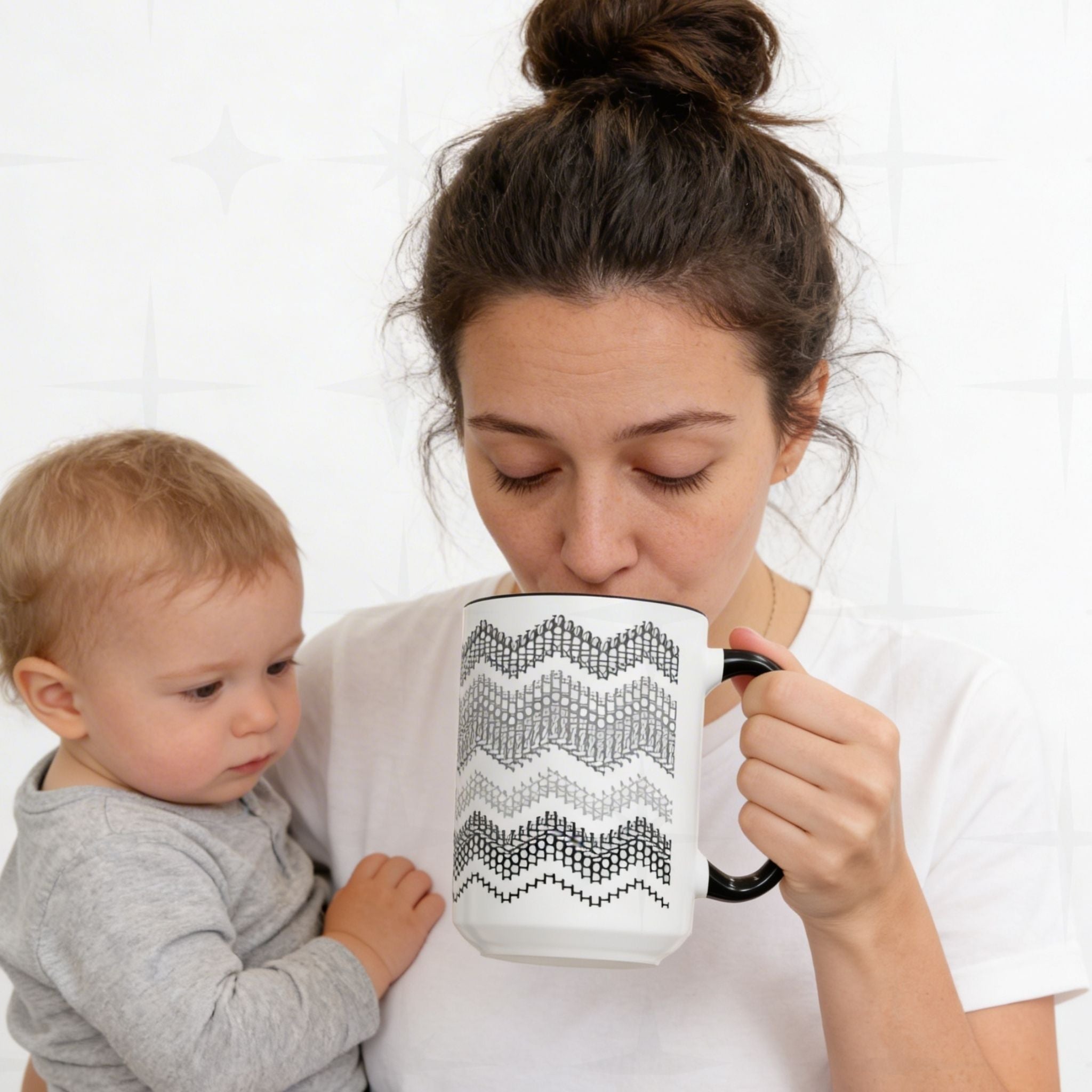 Mother holding a baby on hip while drinking from Gray & Black hidden message mug 15oz — white tile background lifestyle shot, mug showing charcoal-to-black typographic wave pattern