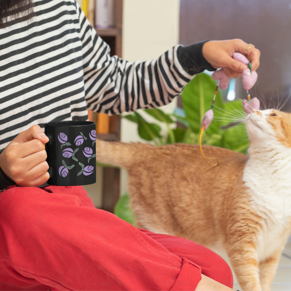 Person in striped shirt holding lilac purple hidden message political mug while playing with an orange and white cat, floral protest design on black ceramic