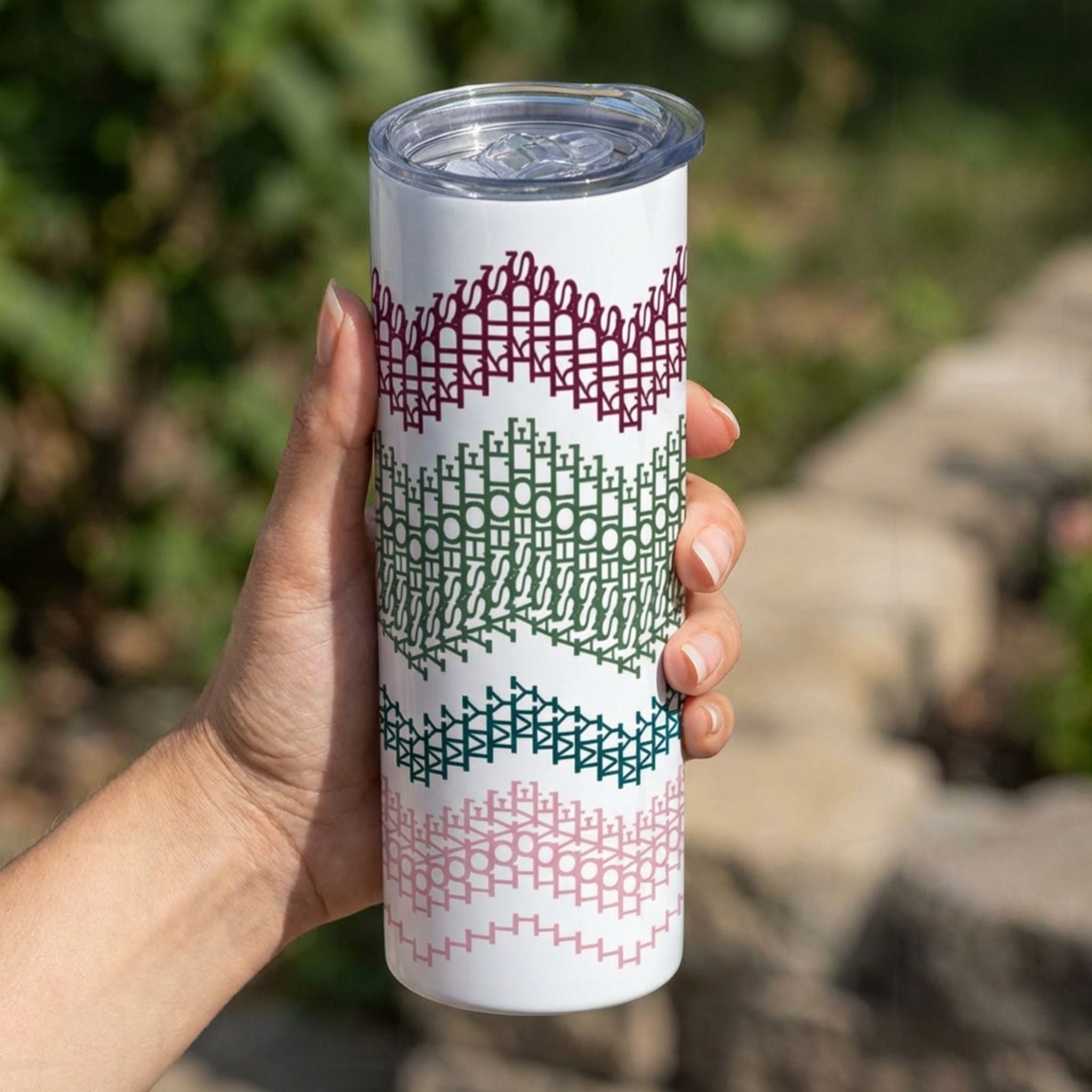 Hand holding a 20 oz skinny stainless steel tumbler with hidden message embroidery stripe design in preppy colorway — burgundy, sage green, forest green, and blush pink chevron bands wrapping the white tumbler body