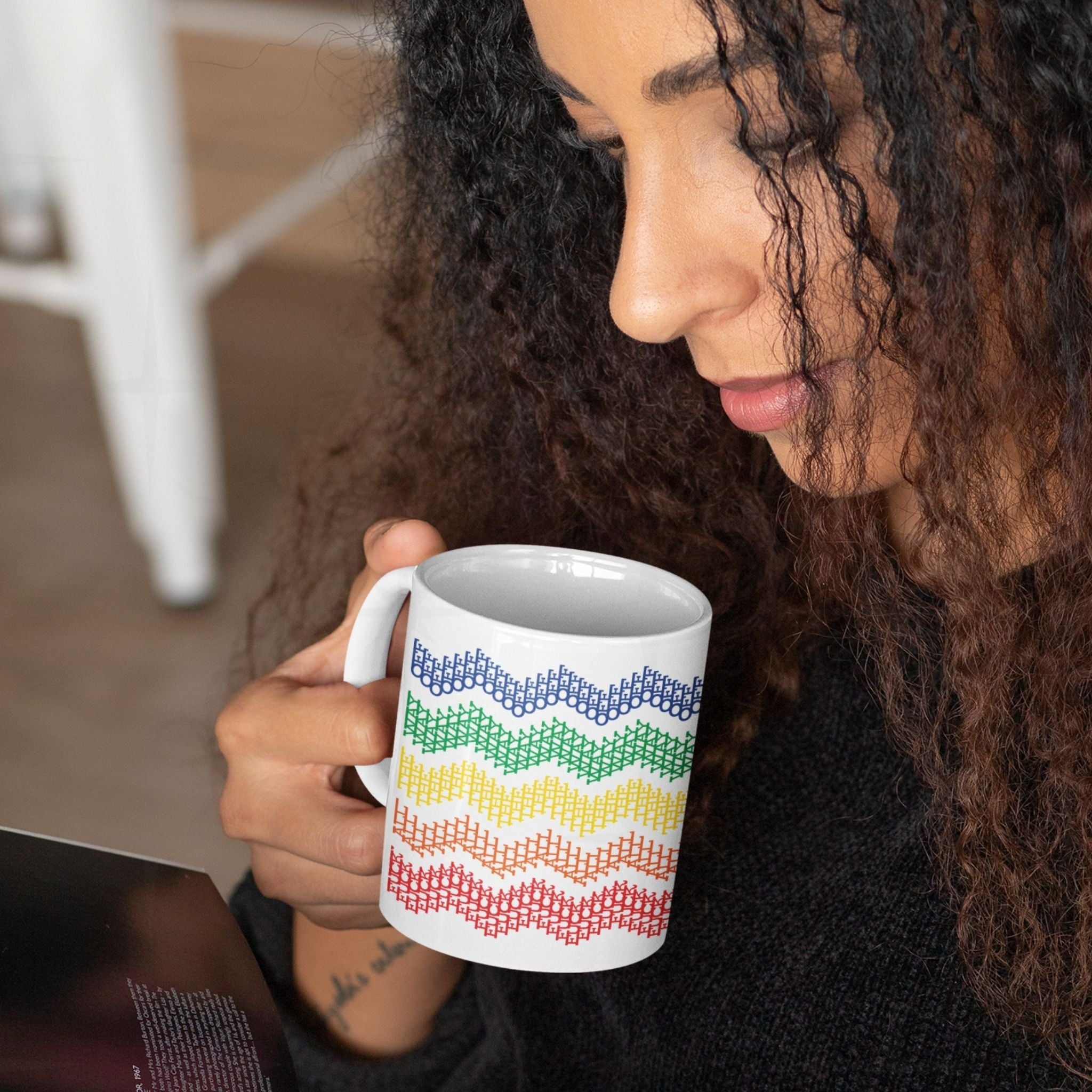 Woman with curly hair reading while holding rainbow stripes hidden profanity chevron mug, wrist tattoo visible