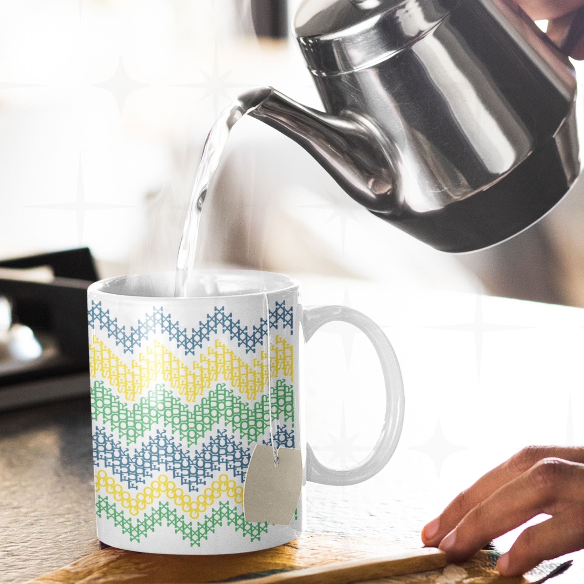 Hidden message cross stitch mug with Go Fuck Yourself text visible in chevron pattern, tea bag inside, hot water being poured from kettle